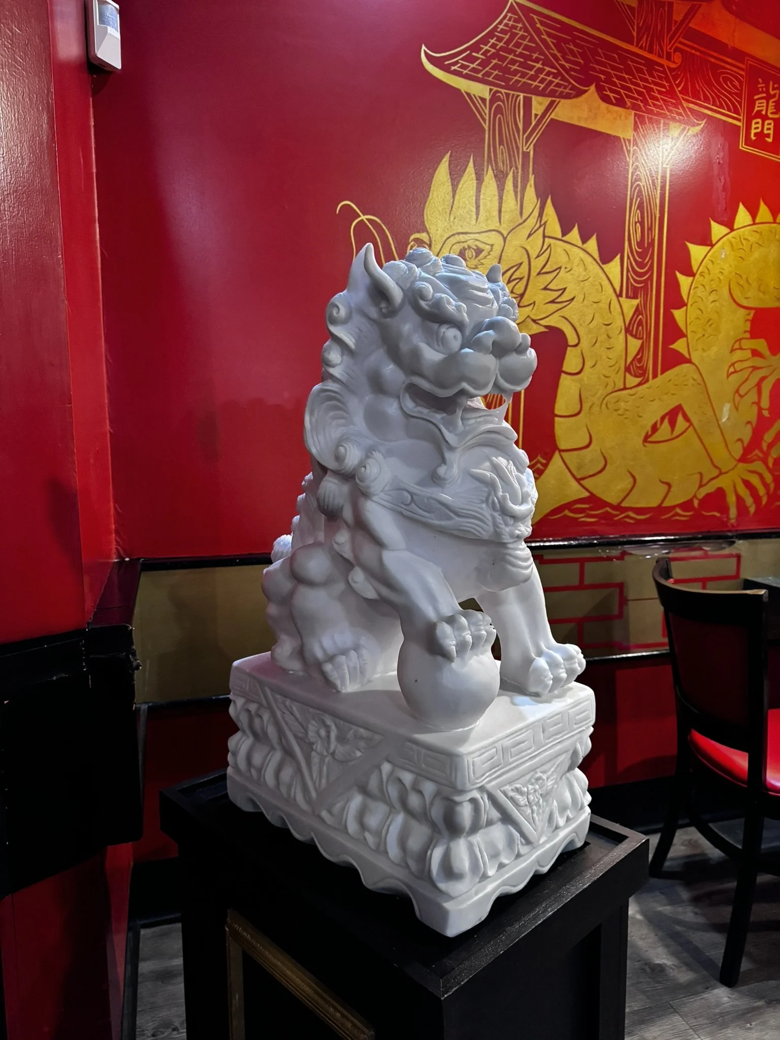 White Chinese guardian lion sculpture on a black stand against a red wall with gold Chinese characters and designs.