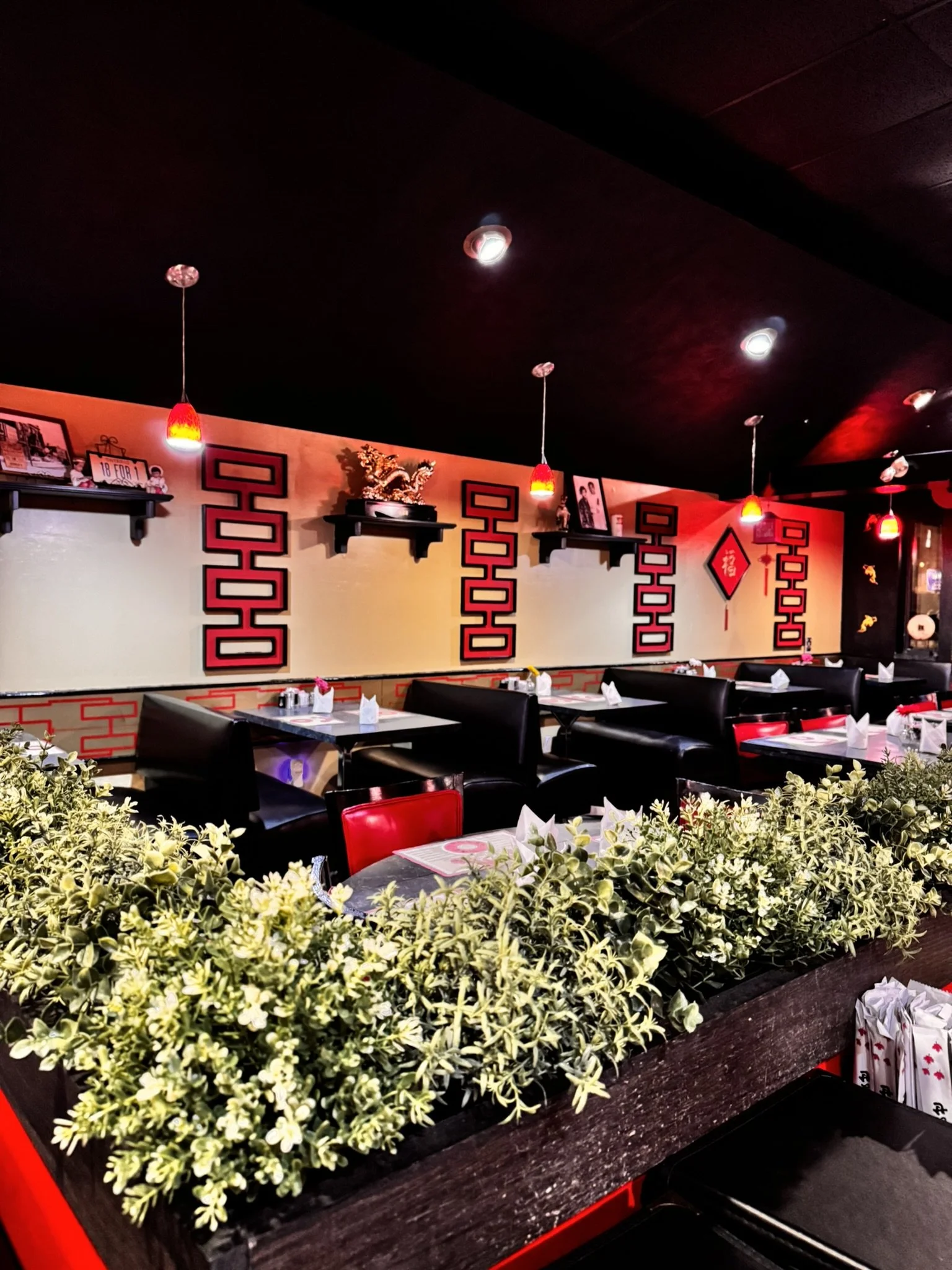 A restaurant interior with a bar counter in the foreground surrounded by a green plant border. The wall behind has decorative shelves with framed photos, a ceramic dragon, and a red Chinese character decoration. Red pendant lights hang from the dark 
