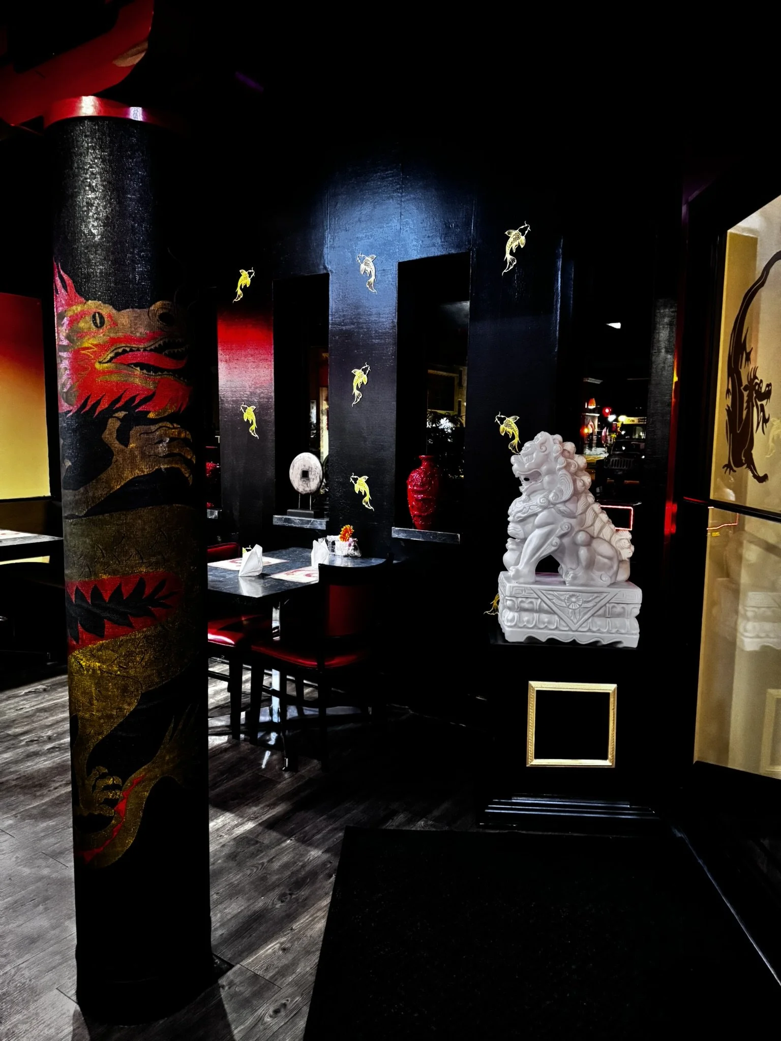 Interior of a restaurant or bar with dark walls decorated with Asian artwork, including a dragon and koi fish stickers, and a white stone lion statue on a black pedestal.
