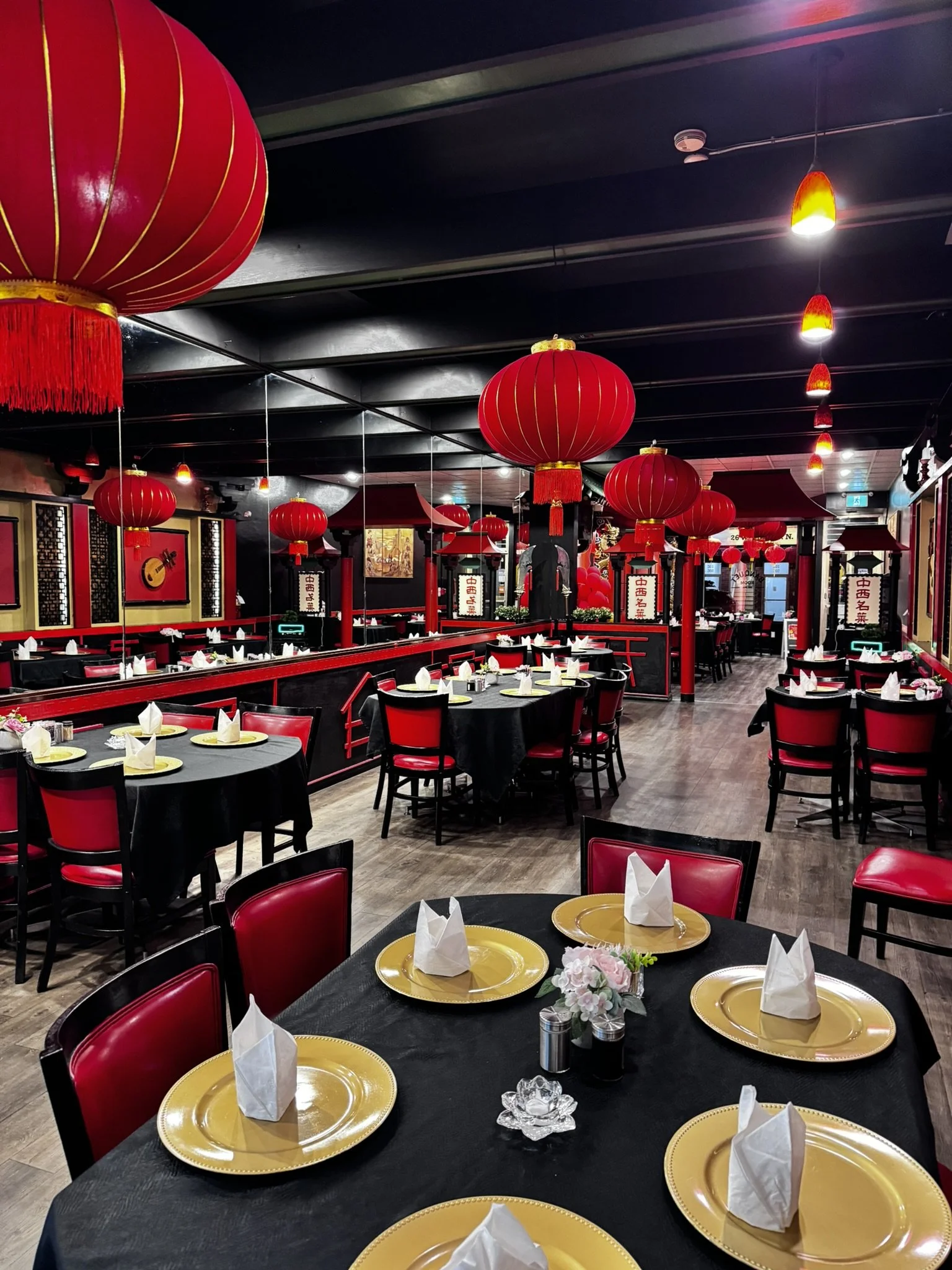 A restaurant decorated for a celebration, with red Chinese lanterns hanging from the ceiling, black tables with yellow plates and white napkins, floral centerpieces, and Chinese-themed wall art.