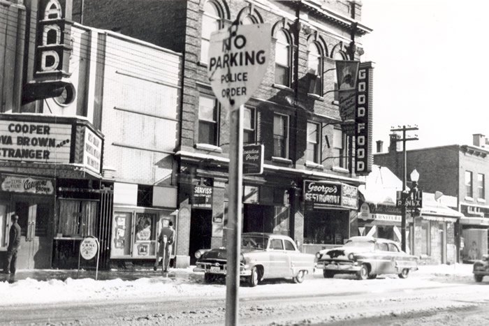 Street scene in a historic American city with vintage cars parked along the snow-covered road, buildings with signs for Good Food and George's Steakhouse, and a pole with a no parking police order sign.