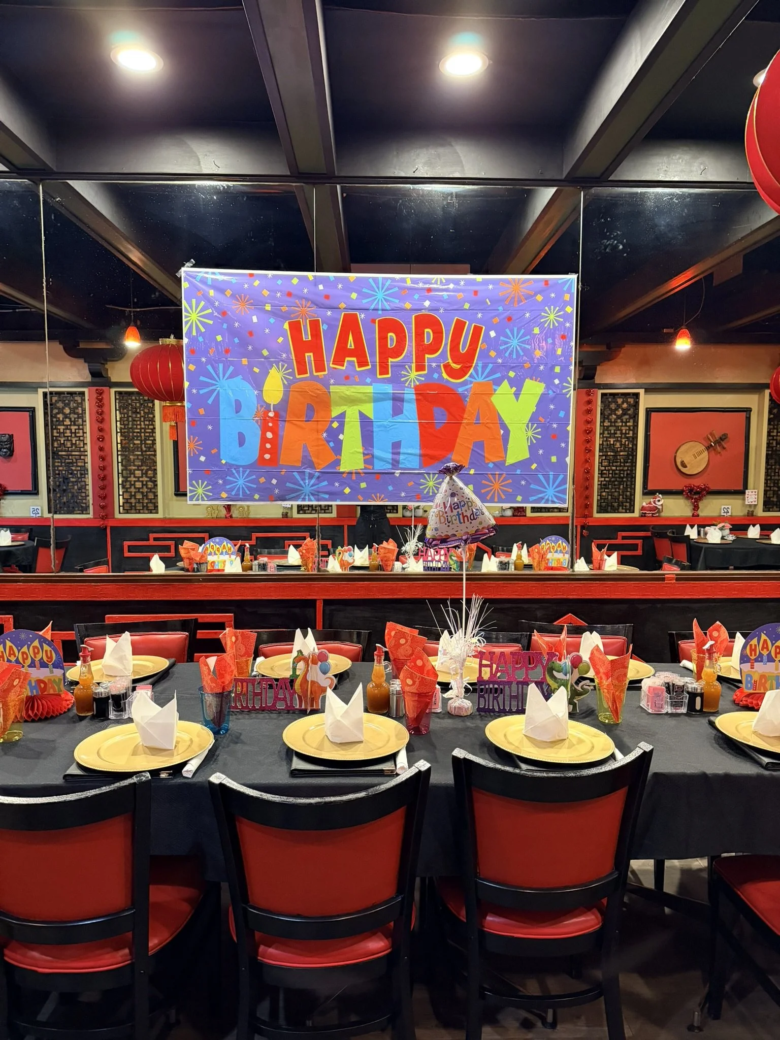 Decorated party room with a large colorful sign saying 'Happy Birthday' on a mirror wall, tables set with birthday hats, napkins, paper plates, and tea bottles, and red and black chairs surrounding the tables.