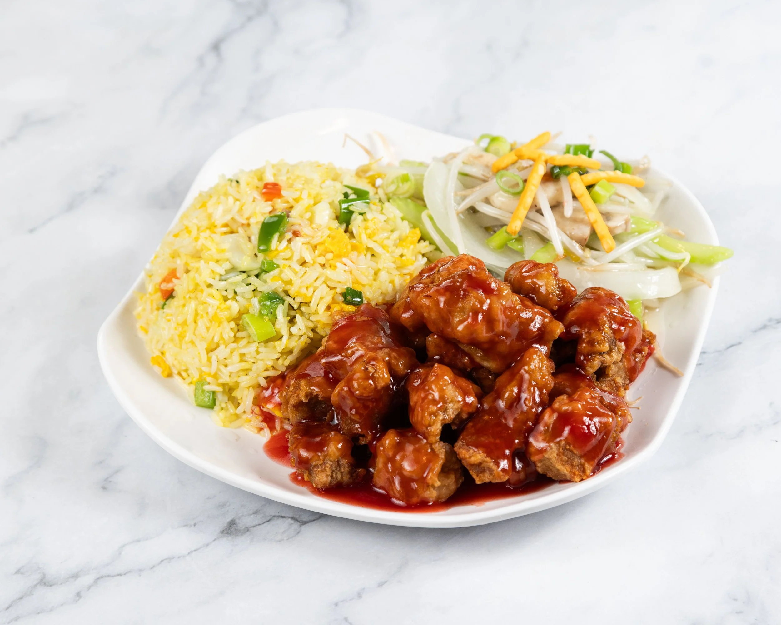 A white bowl containing fried rice with vegetables, a serving of stir-fried noodles with vegetables, and meatballs in a red sauce, placed on a white marble surface.
