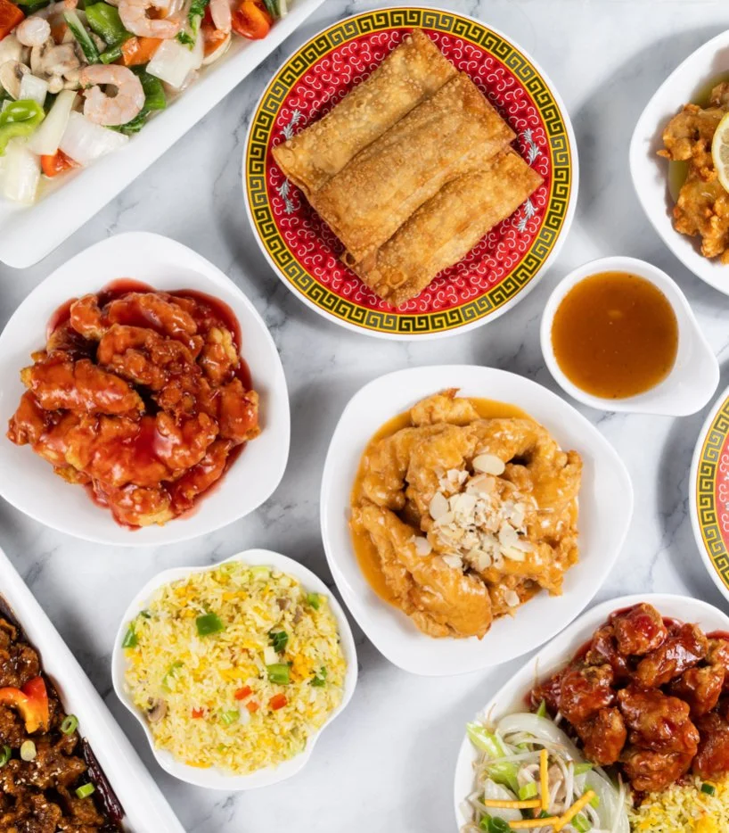 Various Chinese dishes on white serving plates, including spring rolls, sweet and sour chicken, fried chicken with almonds, pineapple fried rice, orange chicken, and stir-fried beef with vegetables.