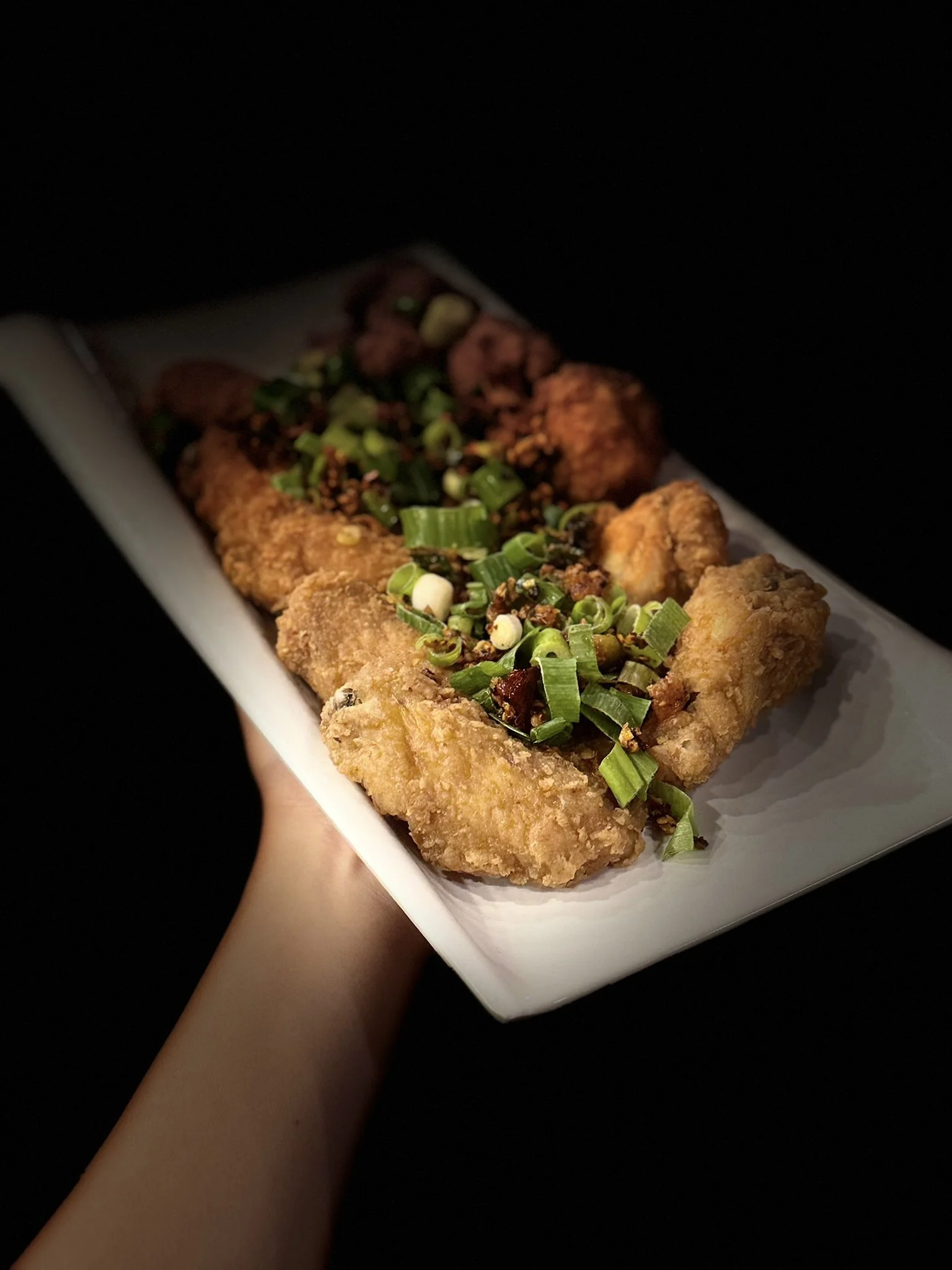 Bite-sized fried chicken pieces garnished with chopped green onions on a white rectangular plate held by a person against a black background.
