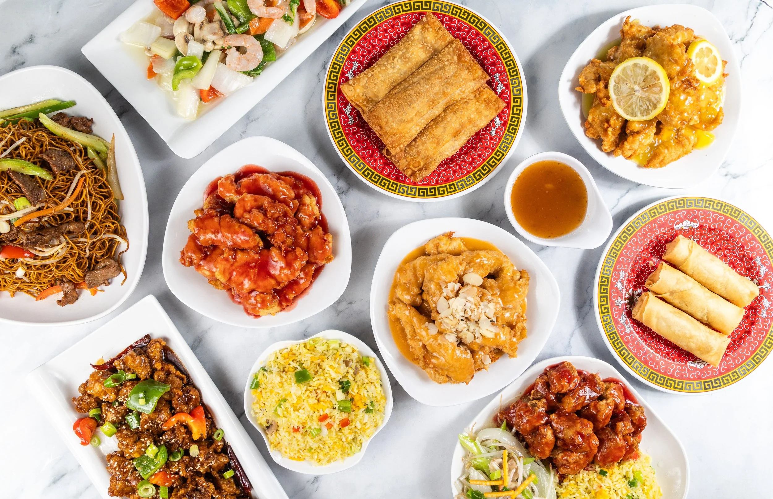 A variety of Chinese dishes on white plates and bowls, including fried spring rolls, sweet and sour chicken, fried rice, chow mein, and vegetable stir-fry, arranged on a marble surface.
