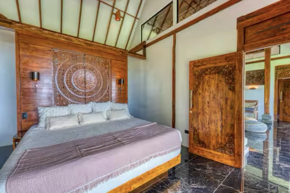 A bedroom with wooden walls and a vaulted ceiling, featuring a large bed with white pillows and a beige bedspread. There is ornate woodwork on the headboard and door, with dark marble flooring.