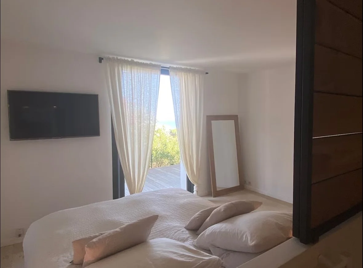 A bedroom with a large glass sliding door leading to a balcony with outdoor seating and a view of lush green trees.