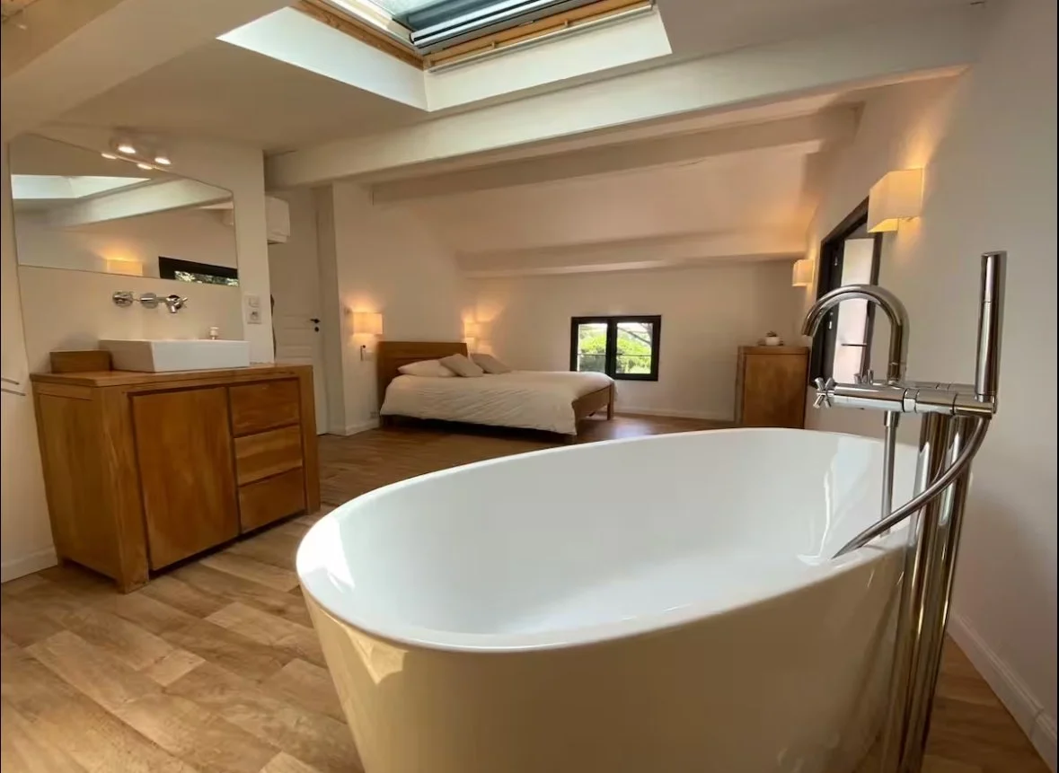 A bedroom with wooden walls and a vaulted ceiling, featuring a large bed with white pillows and a beige bedspread. There is ornate woodwork on the headboard and door, with dark marble flooring.