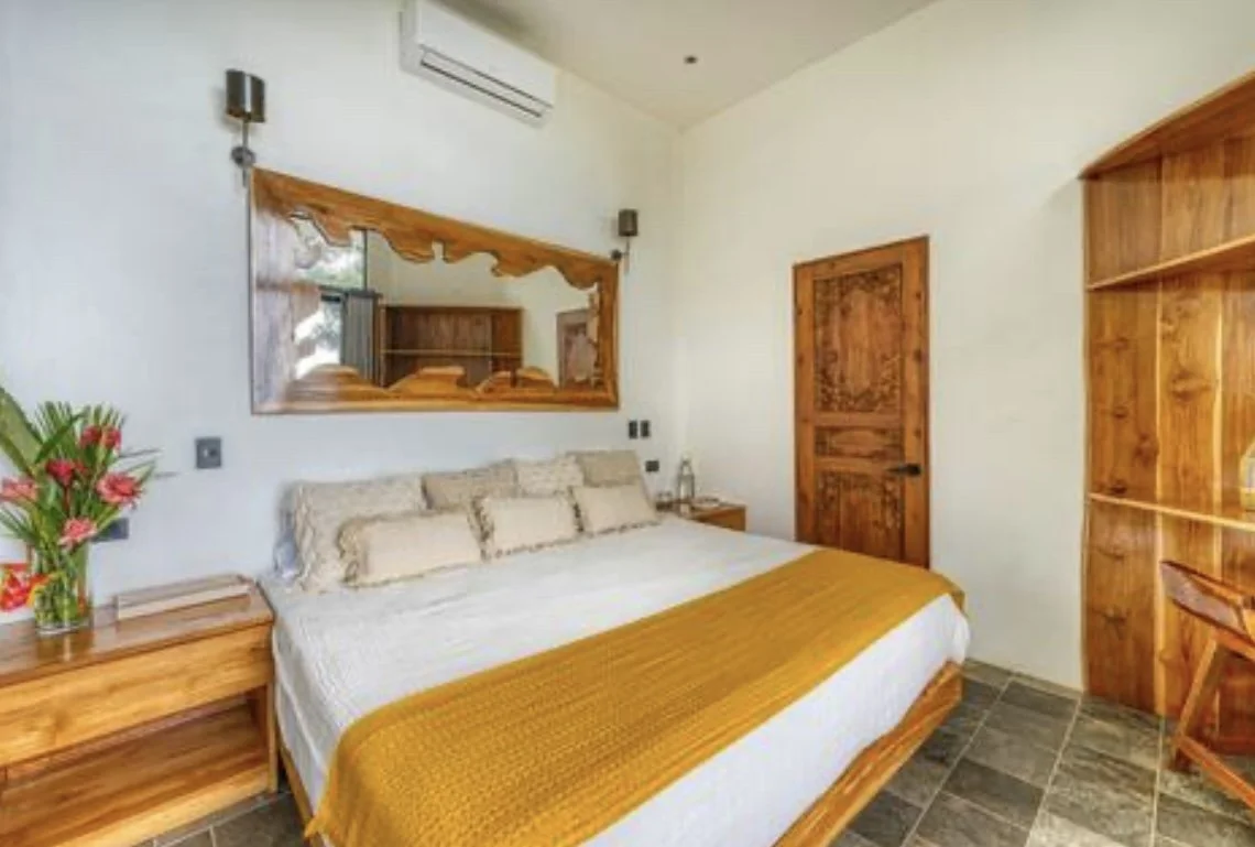 A bedroom with a large bed, wooden furniture, a mirror, and a sliding door, decorated in neutral colors with a mustard-colored throw.