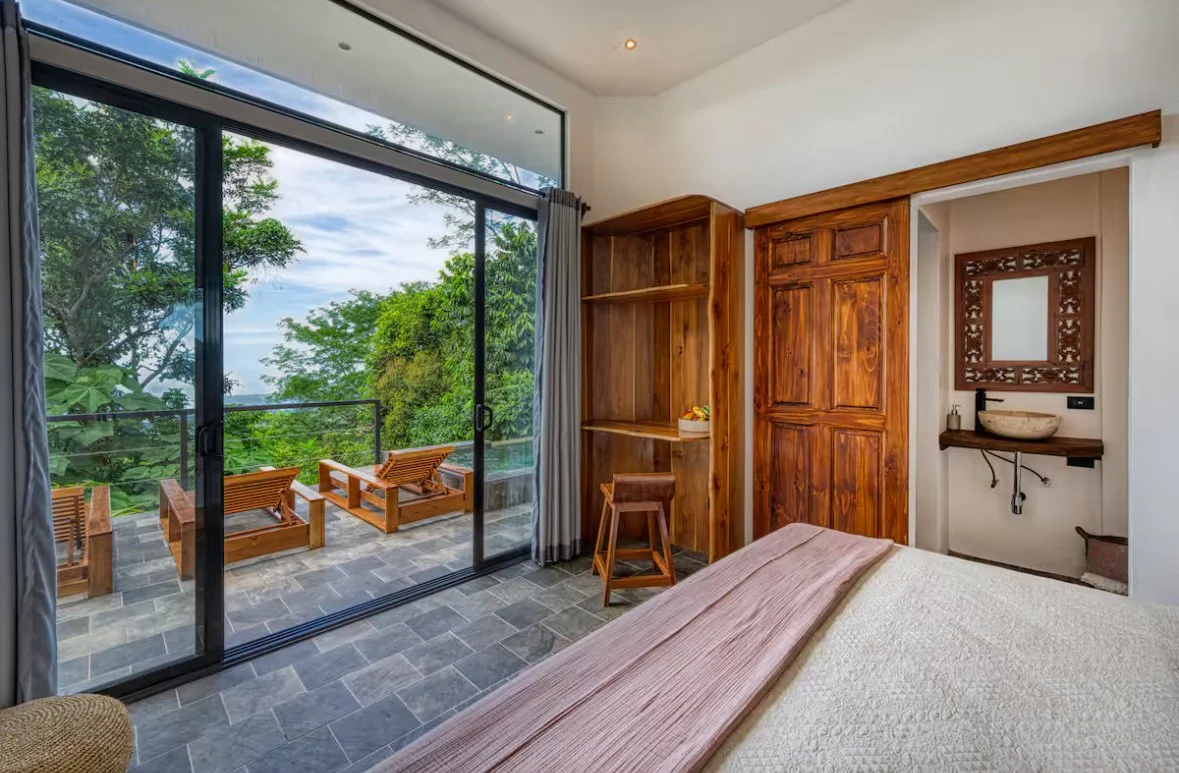A bedroom with a large glass sliding door leading to a balcony with outdoor seating and a view of lush green trees.