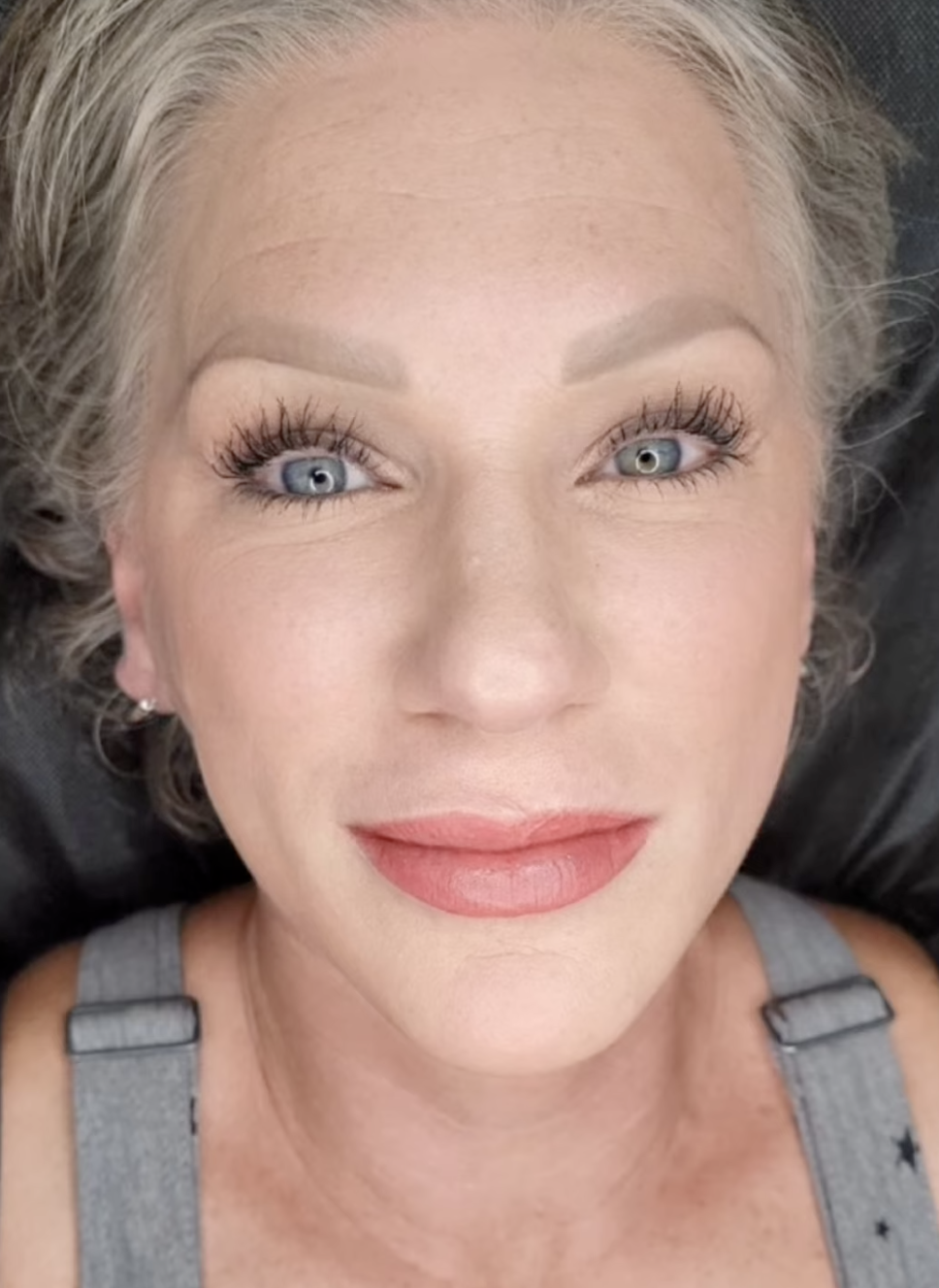 Close-up of a woman with blue eyes, blonde hair, and light makeup, lying down on a black surface, wearing a gray top with black star patterns.