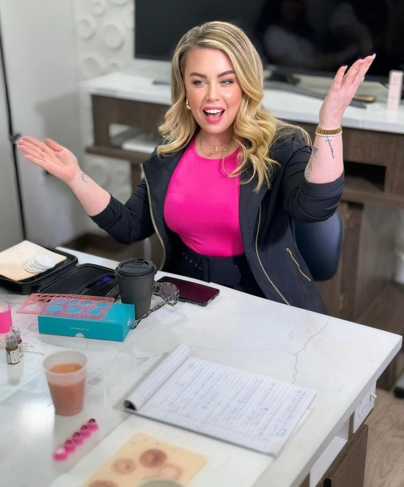 A woman with blonde hair, wearing a black jacket and pink top, sitting at a white table with makeup supplies and notes, smiling and gesturing with both hands.