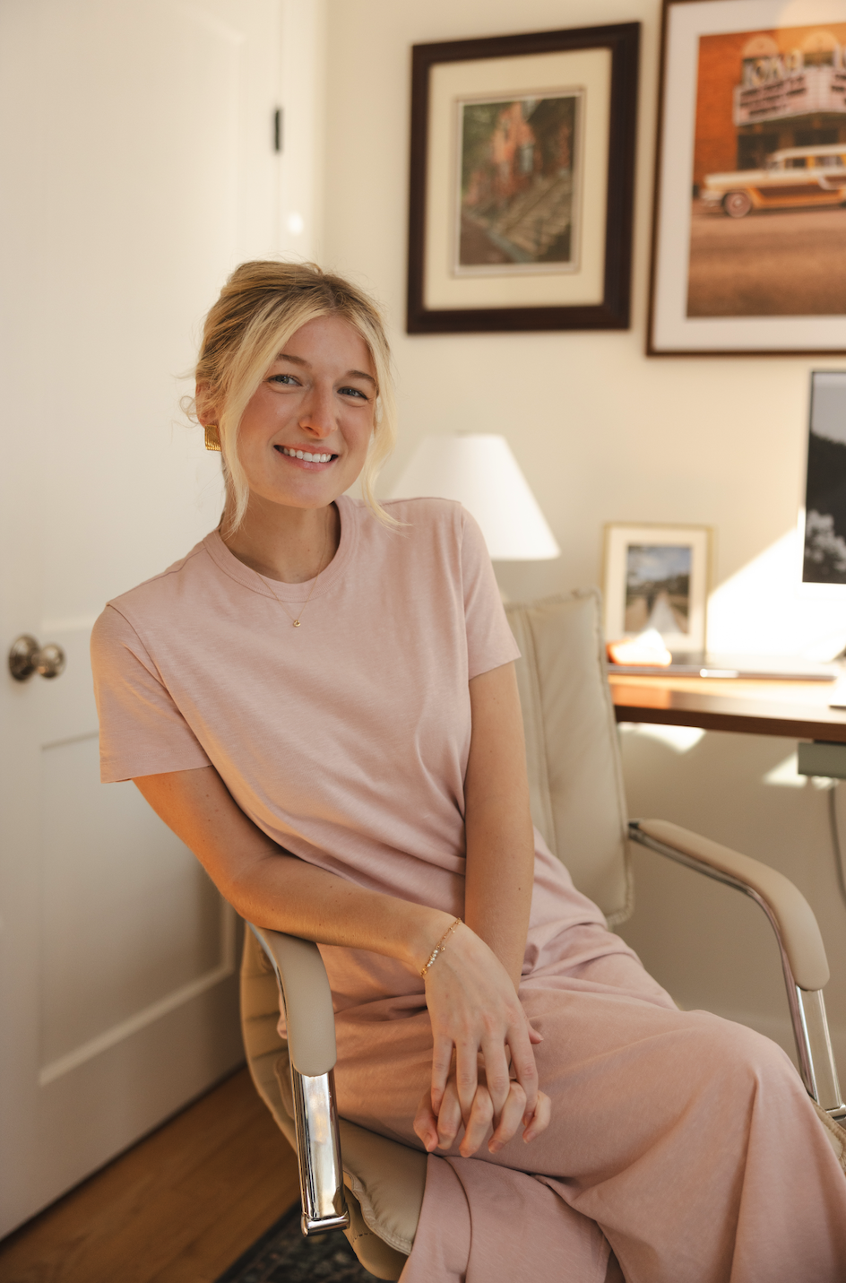 A young woman with blonde hair sitting on a beige chair in a cozy room, smiling at the camera. She is wearing a light pink t-shirt and matching pants, accessorized with gold jewelry. Behind her, framed pictures hang on a cream-colored wall, with a desk, a laptop, a lamp, and some small decor items in the background.