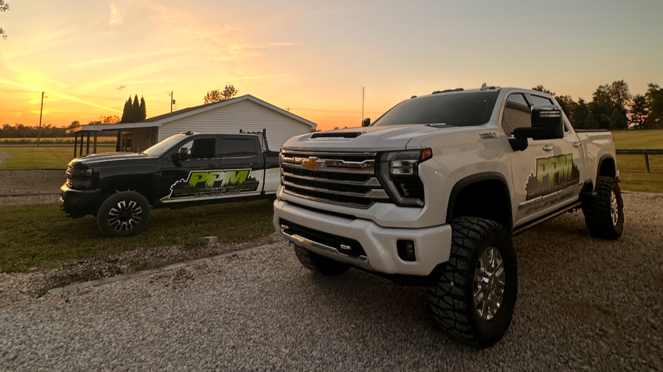 Two pickup trucks parked on grass and gravel in front of a small white building during sunset. The trucks have a logo with the letters 'PFM' on their sides.