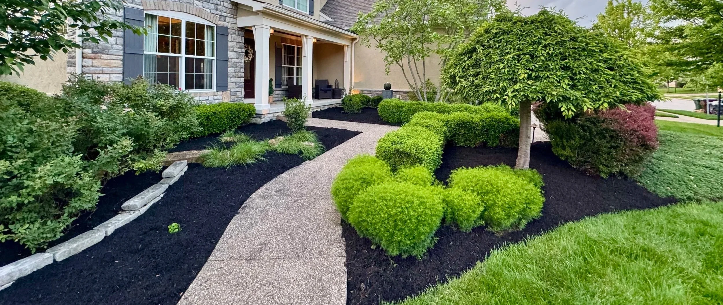 Front yard landscaped garden with a curved walkway, small shrubs, and trees in front of a house with stone and siding exterior.