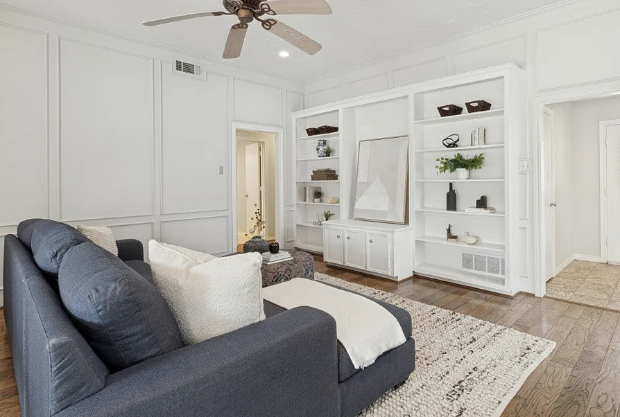Living room with white walls, wooden floor, dark gray sofa with light-colored pillows, a patterned area rug, and built-in white bookshelves with decorative items. There is a ceiling fan and doorway leading to another room.