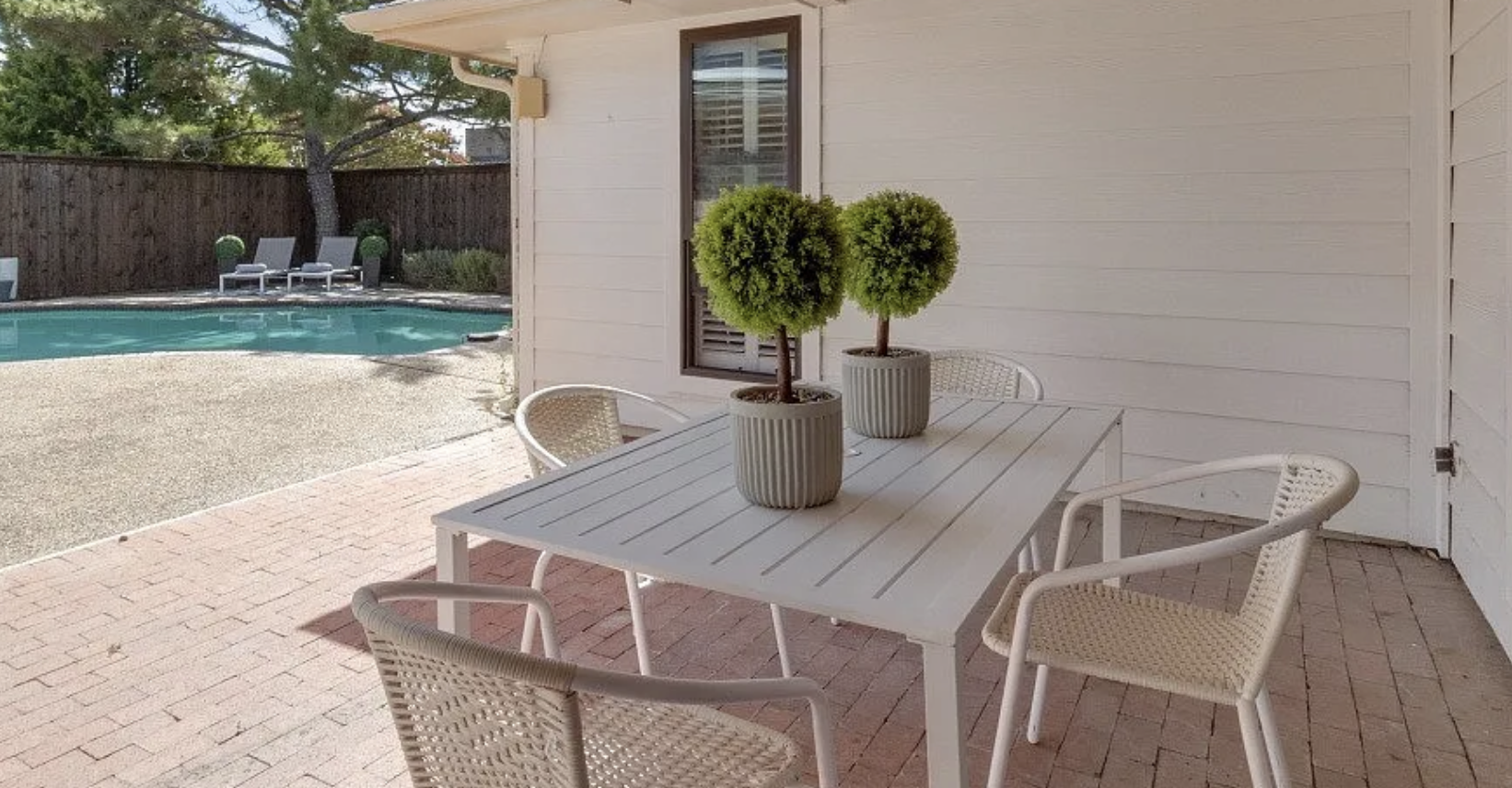 Patio with white table and four wicker chairs, two with potted topiary plants, and a pool with lounge chairs and trees in the background.
