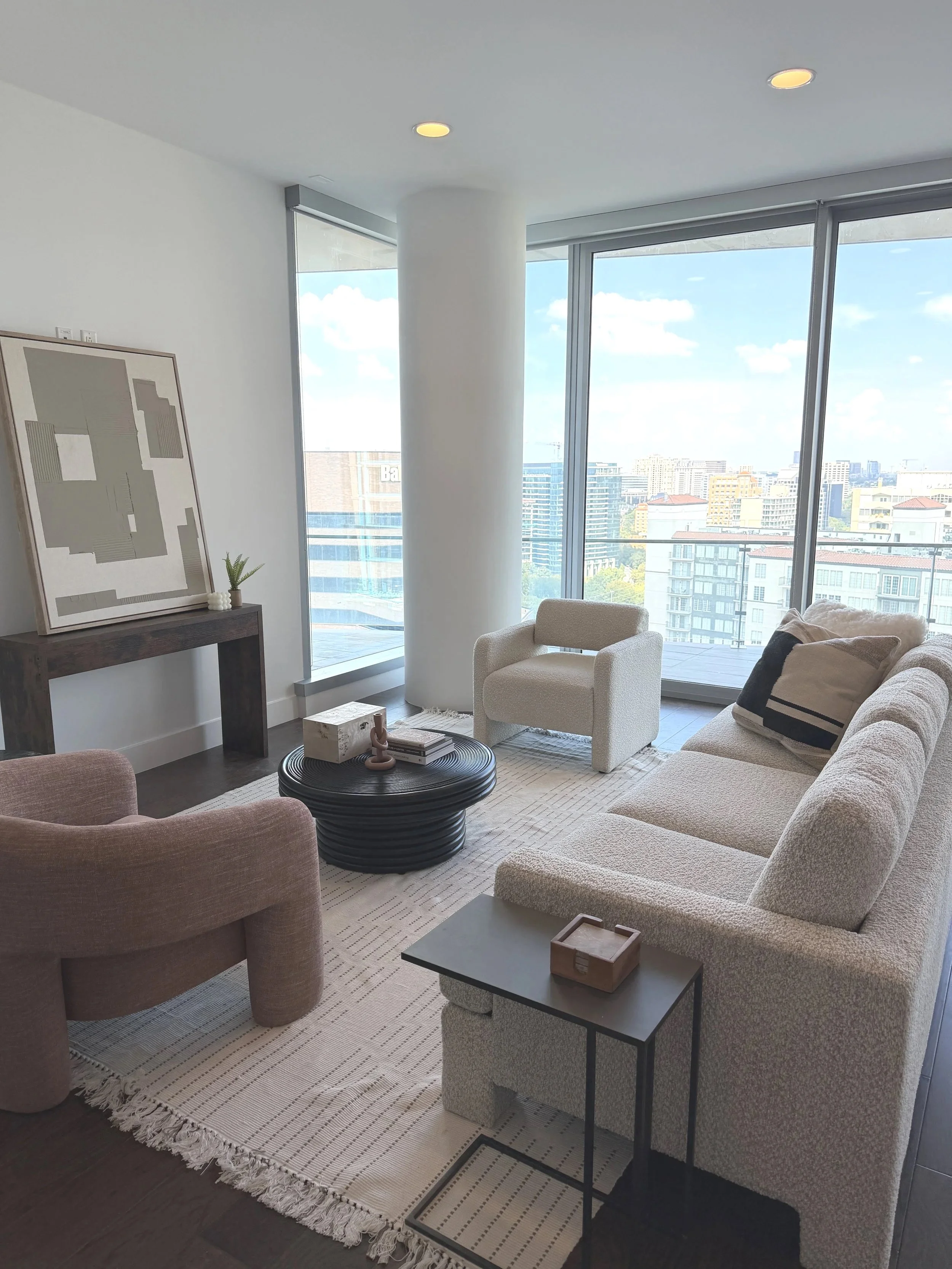 Modern living room with large windows showing city view, white armchair, beige sofa with pillows, black and white striped rug, round black coffee table, framed artwork, and indoor plants.