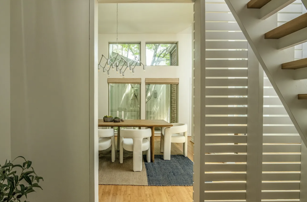 Interior view of a dining area with a wooden table, white chairs, large windows, and a modern chandelier, partially seen through a decorative white slatted room divider.