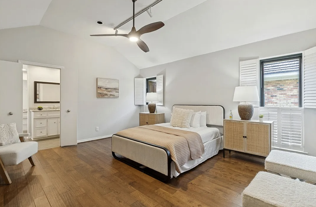 Bright bedroom with white walls, wooden floors, a bed with white bedding, two nightstands with lamps, a chair, and a bathroom visible through a doorway.