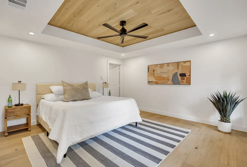 A contemporary bedroom with a white bed, wooden nightstands with lamps, a ceiling fan, and abstract wall art, featuring a striped area rug and a potted plant.