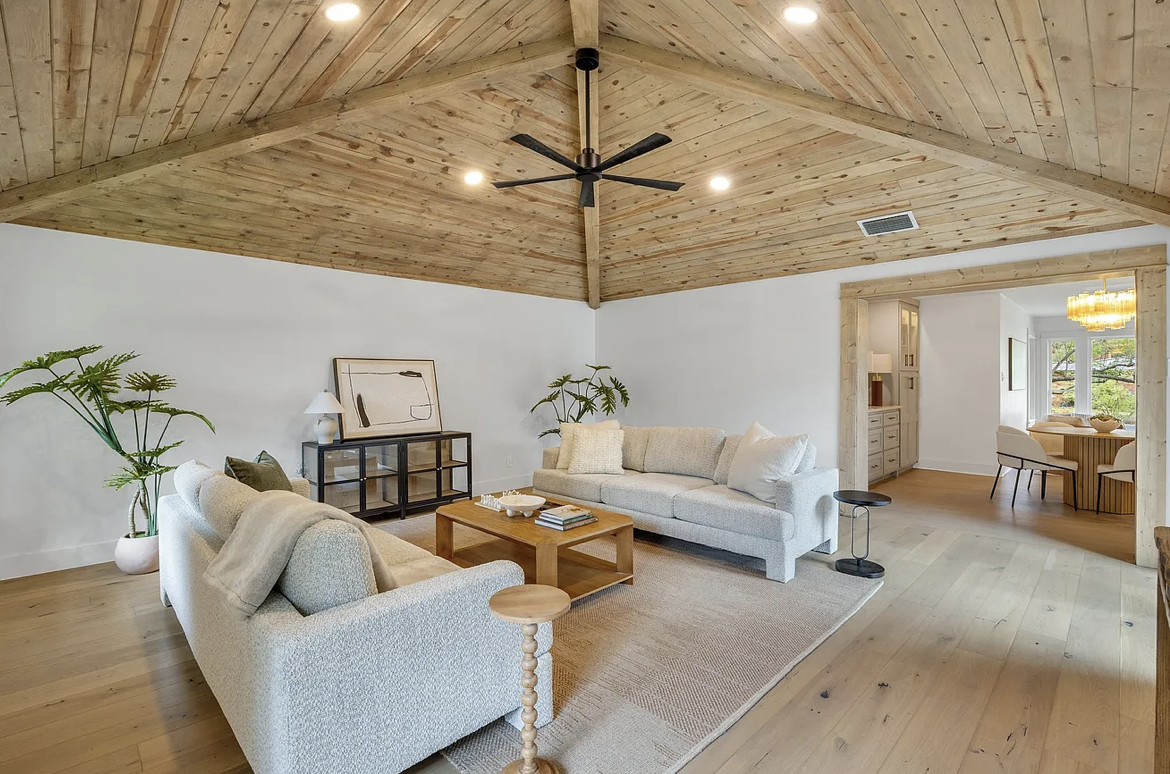 Living room with light-colored sofas, wooden coffee table, large potted plants, a black ceiling fan, wooden ceiling, and hardwood floors, opening to a dining area with a chandelier.
