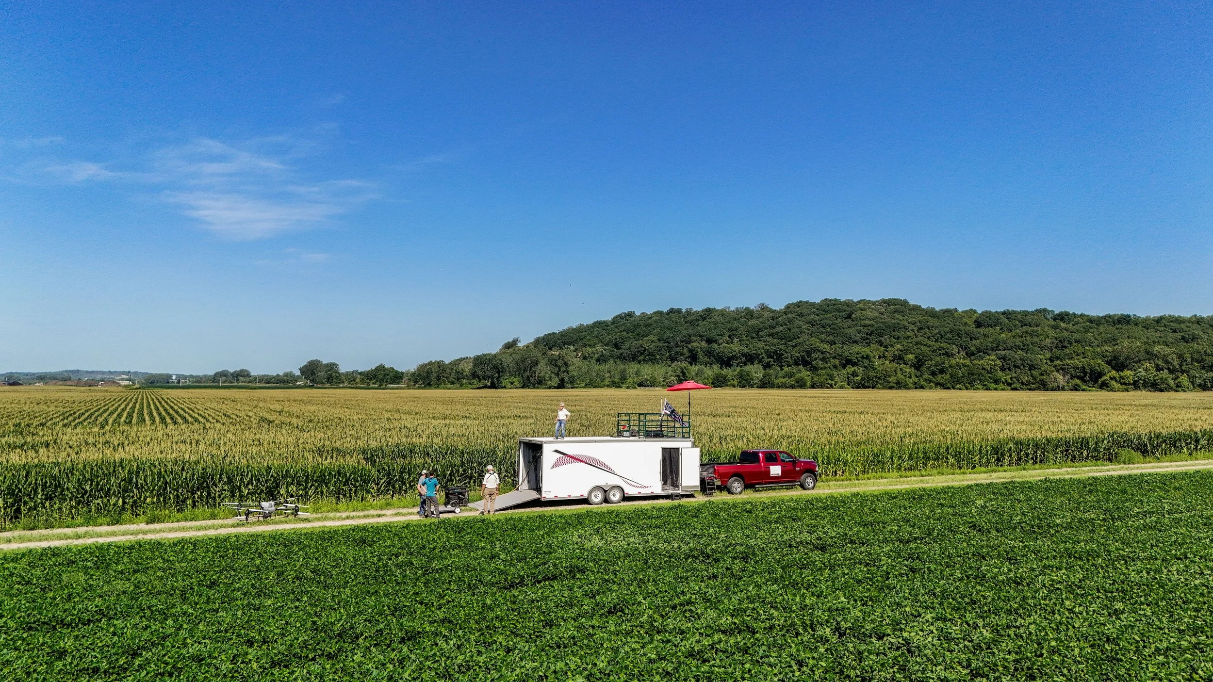 A group of people near a white trailer and a red pickup truck on a farm with rows of crops under a clear blue sky.