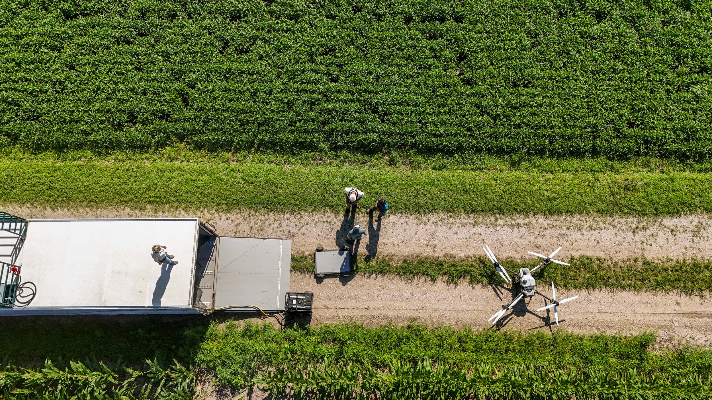 Aerial view of a drone flying along a dirt path next to lush green fields with three people standing on the path, one person on a white trailer, and the drone.