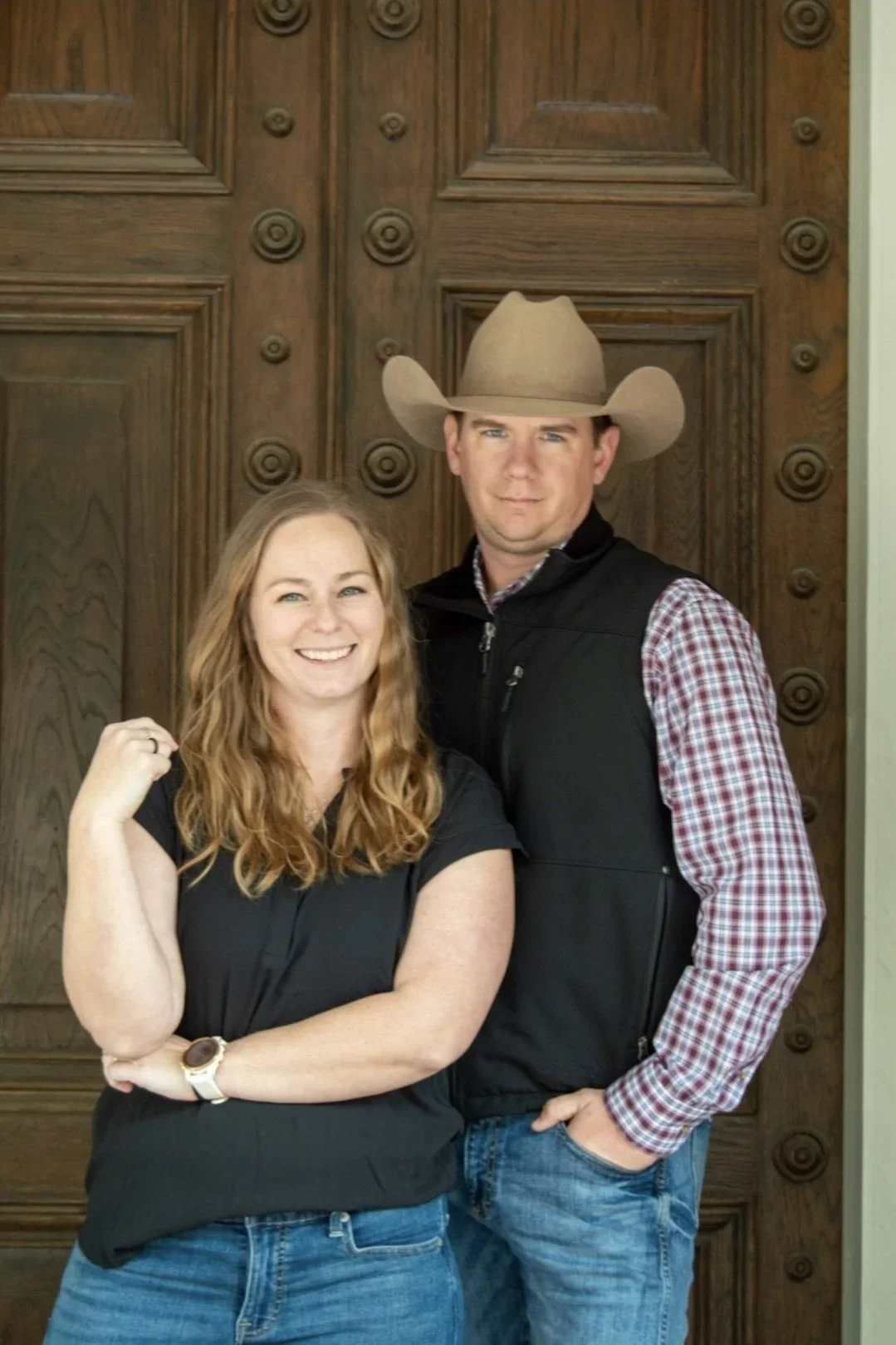 A smiling woman with long, wavy red hair wearing a black shirt and blue jeans, standing with her arm flexed, next to a man in a cowboy hat, plaid shirt, and black vest, with a wooden background.