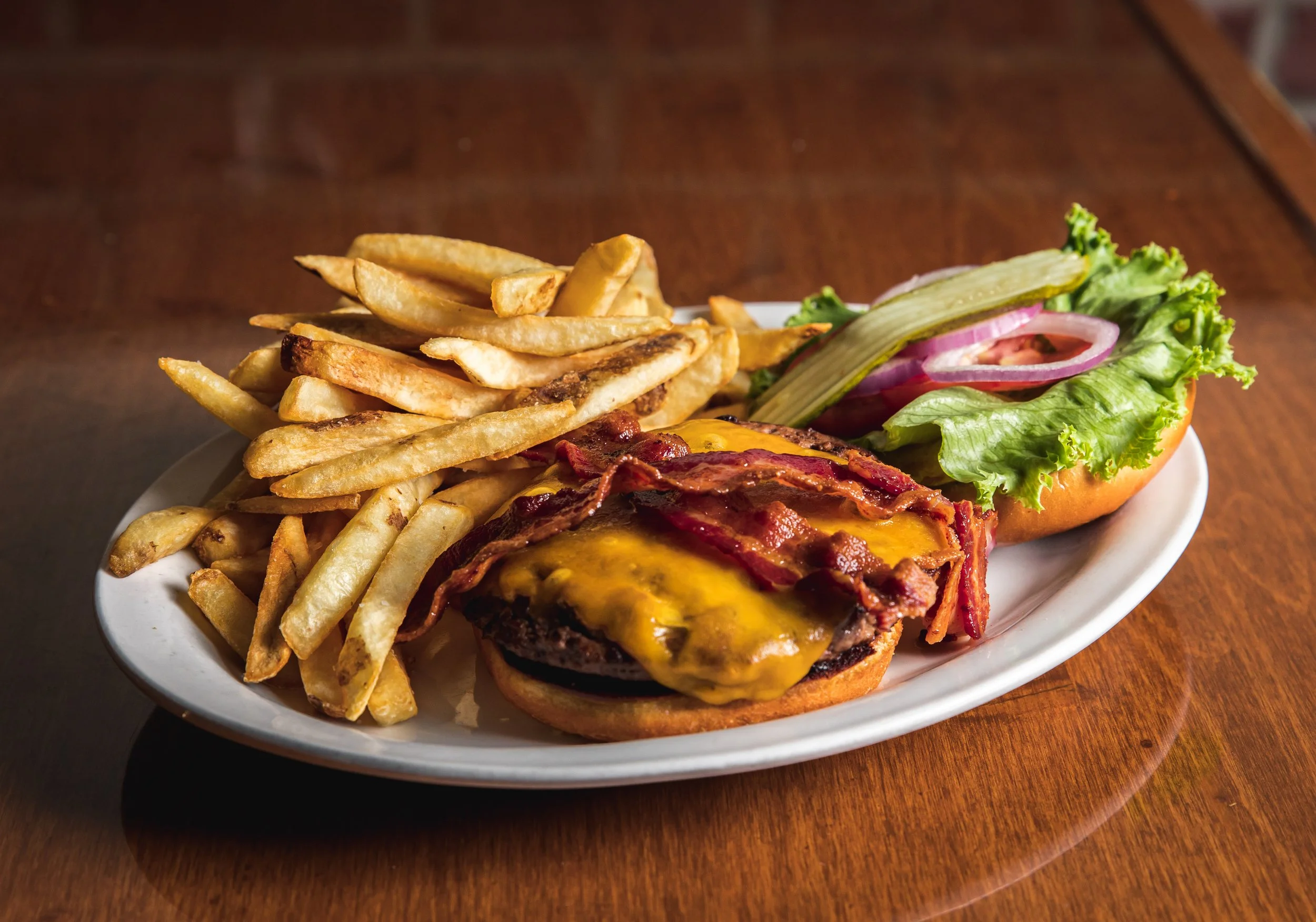 A plate of cheeseburger with bacon, French fries, and a side salad with lettuce, onion, cucumber, and tomato on a wooden table.