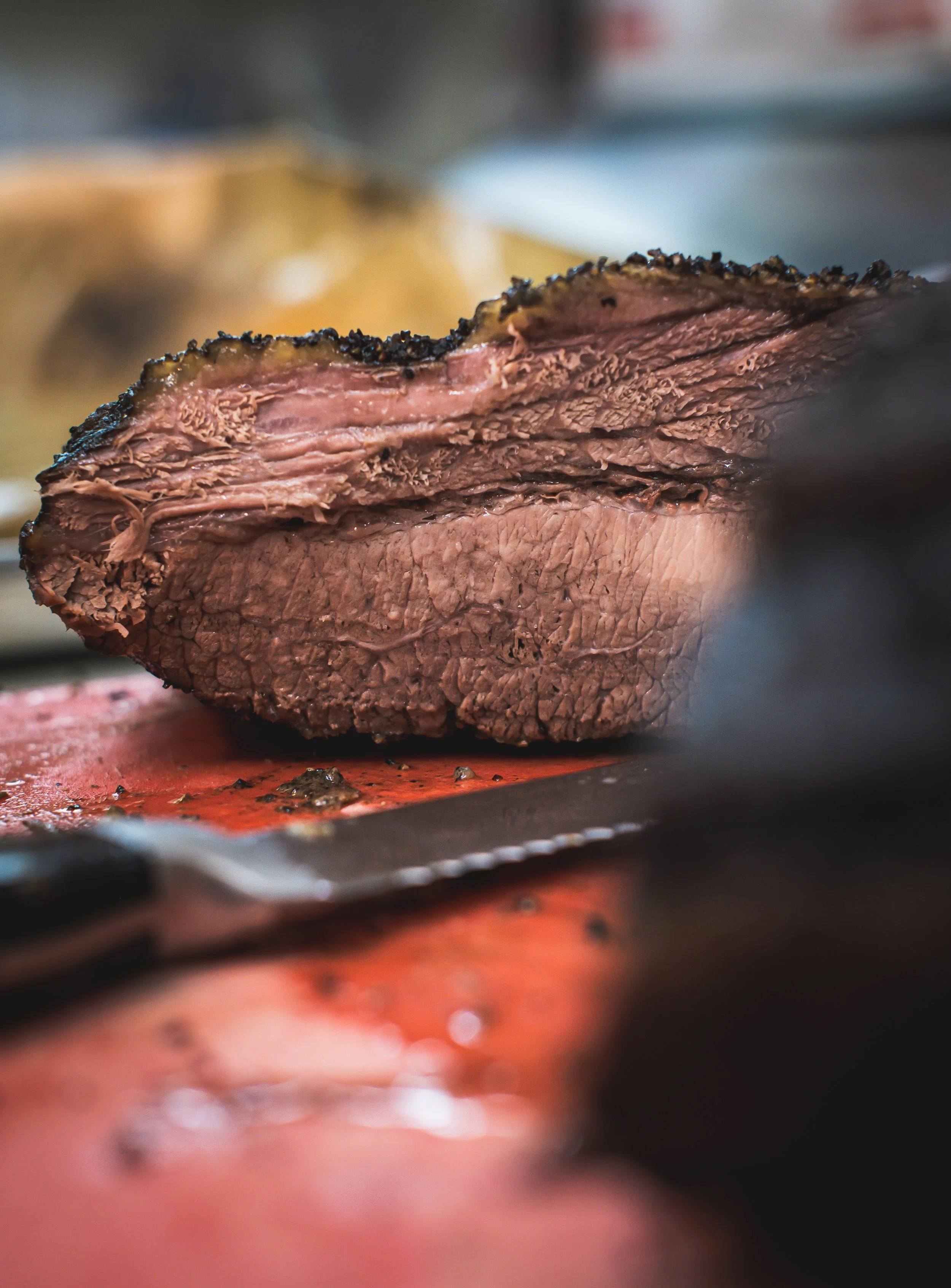 Close-up of a cooked slice of beef brisket with a dark, peppery crust, resting on a cutting board with a carving knife nearby.