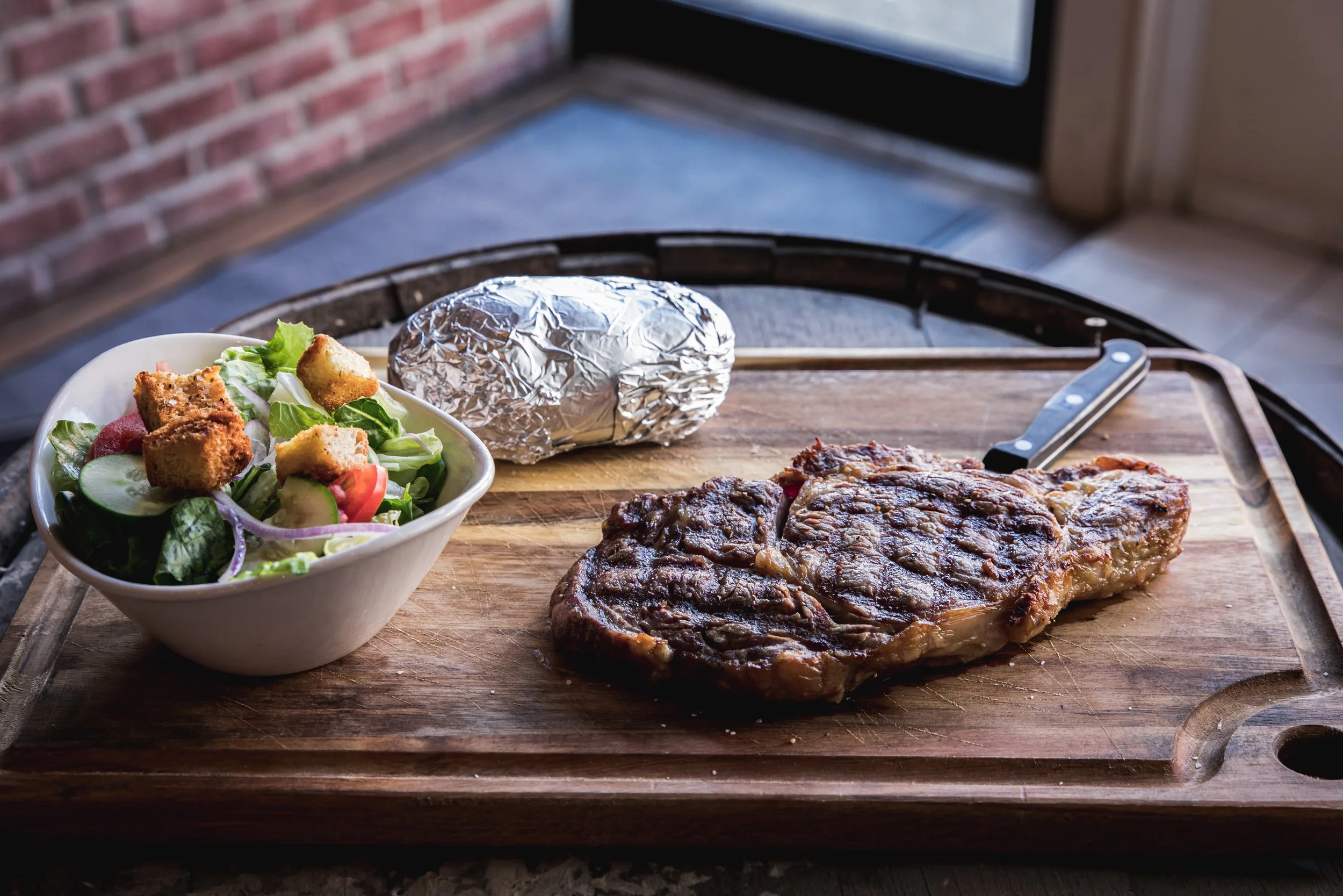 Grilled steak on a wooden cutting board with a side salad in a white bowl and a foil-wrapped baked potato.