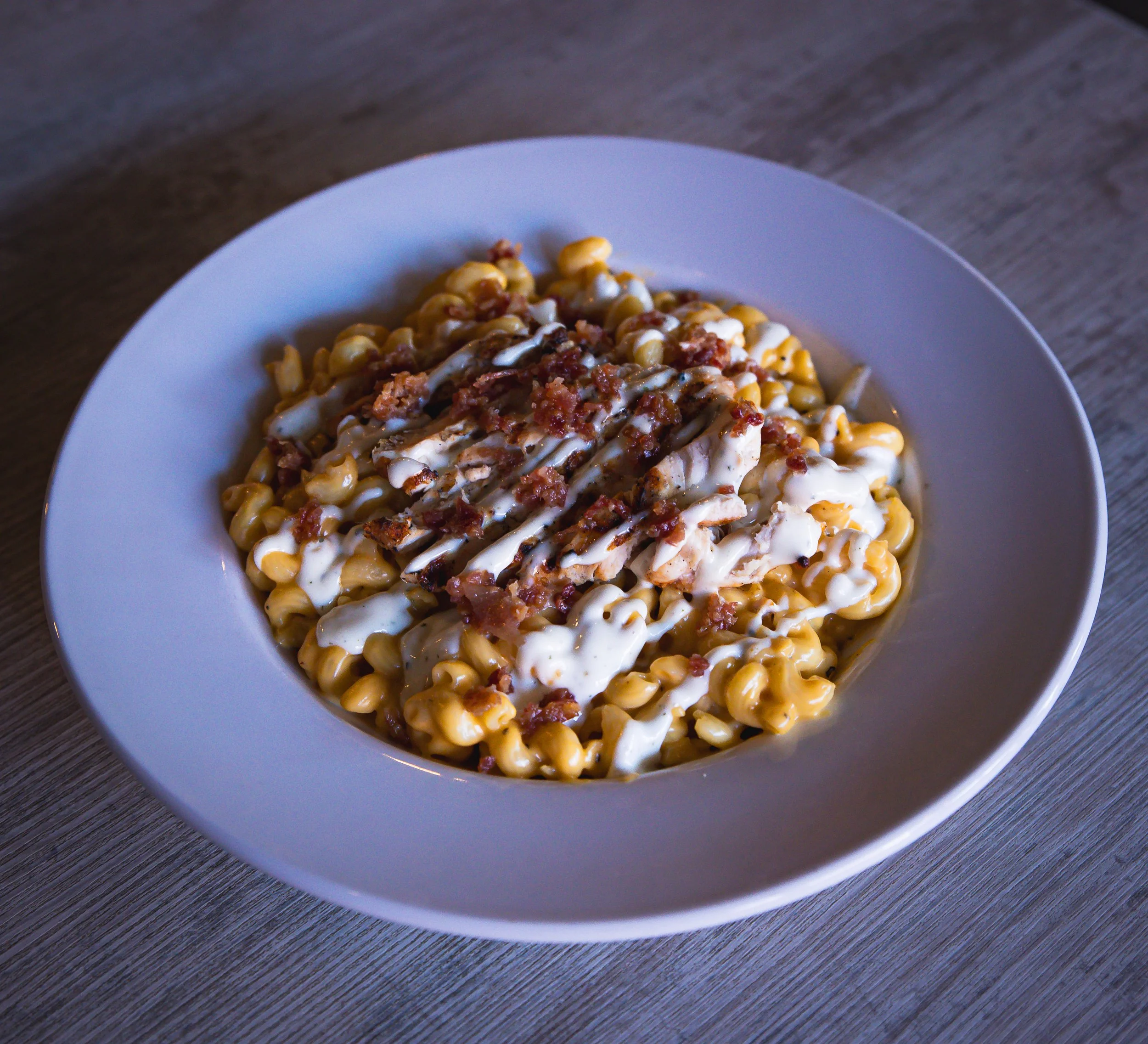 Creamy macaroni and cheese topped with bacon bits and drizzled with white sauce, served in a white bowl on a wooden table.