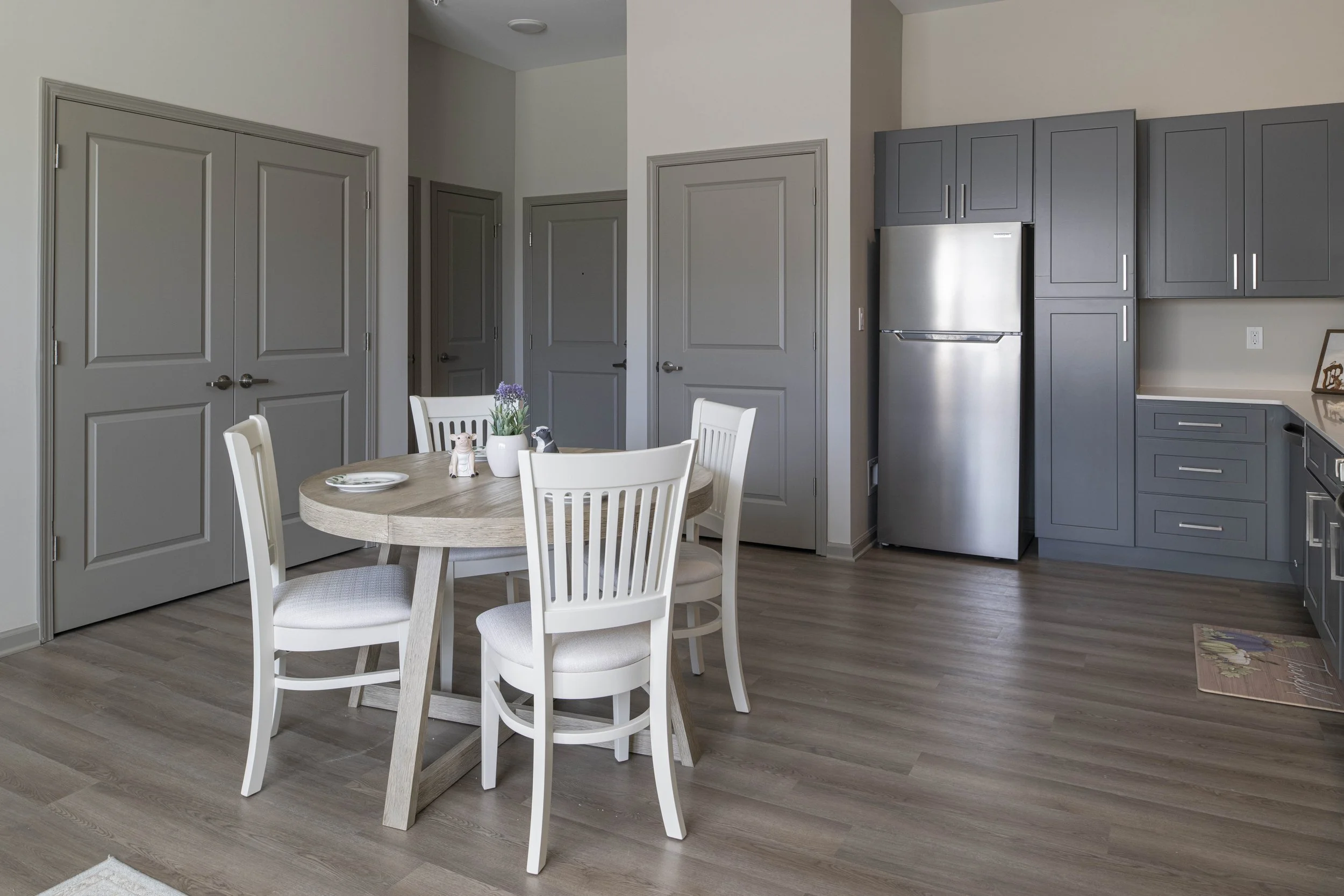 Kitchen with a round dining table and four white chairs, gray cabinets, stainless steel refrigerator, wooden floor, and neutral wall color.
