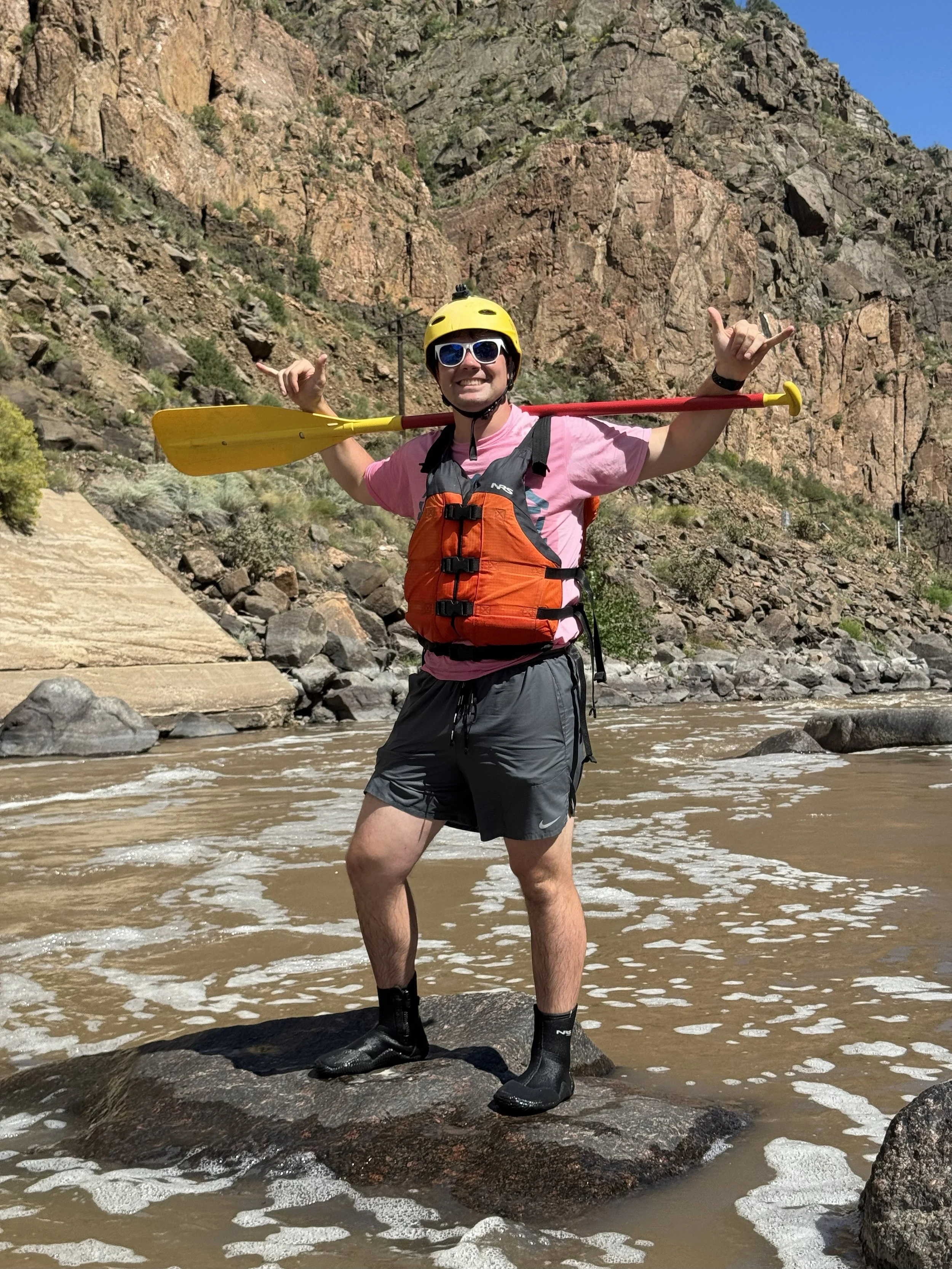 Person in pink shirt and gray shorts standing on a rock in a river, holding a yellow paddle over their shoulders, wearing a yellow helmet, sunglasses, and an orange life jacket, with rocky hills in the background.
