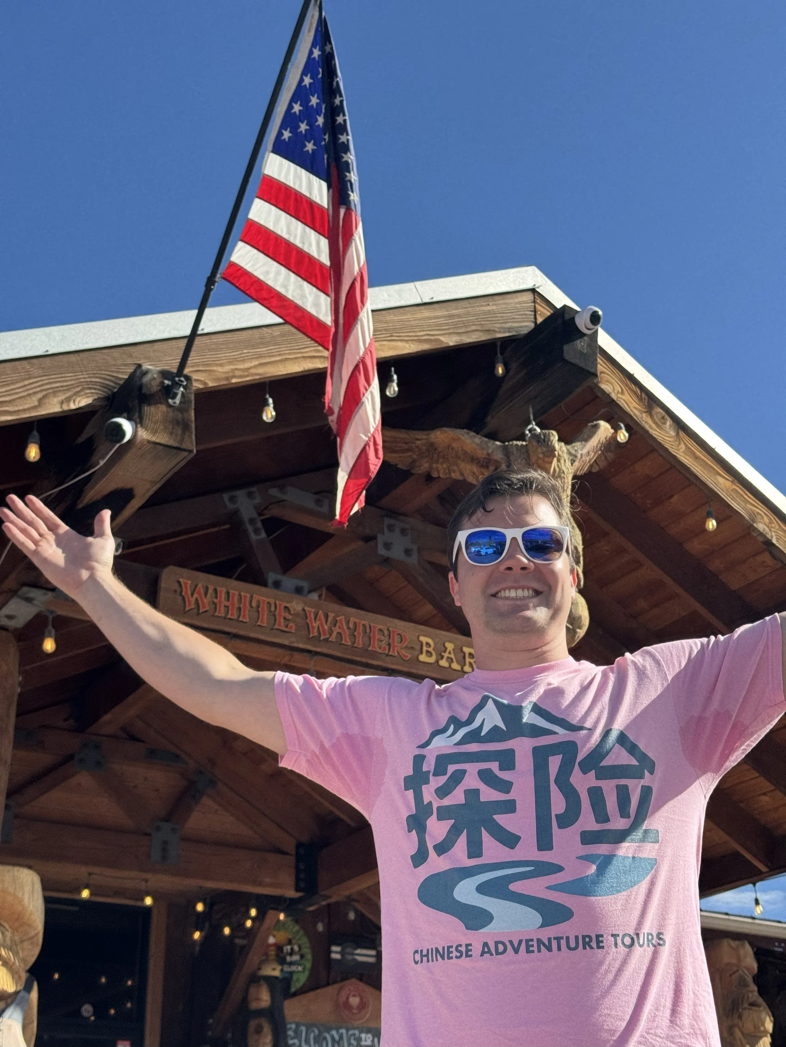 A man wearing sunglasses and a pink T-shirt with Chinese characters and 'Chinese Adventure Tours' text is smiling with arms outstretched in front of a wooden building with an American flag and a sign that reads 'White Water Bar' under a clear blue sky.