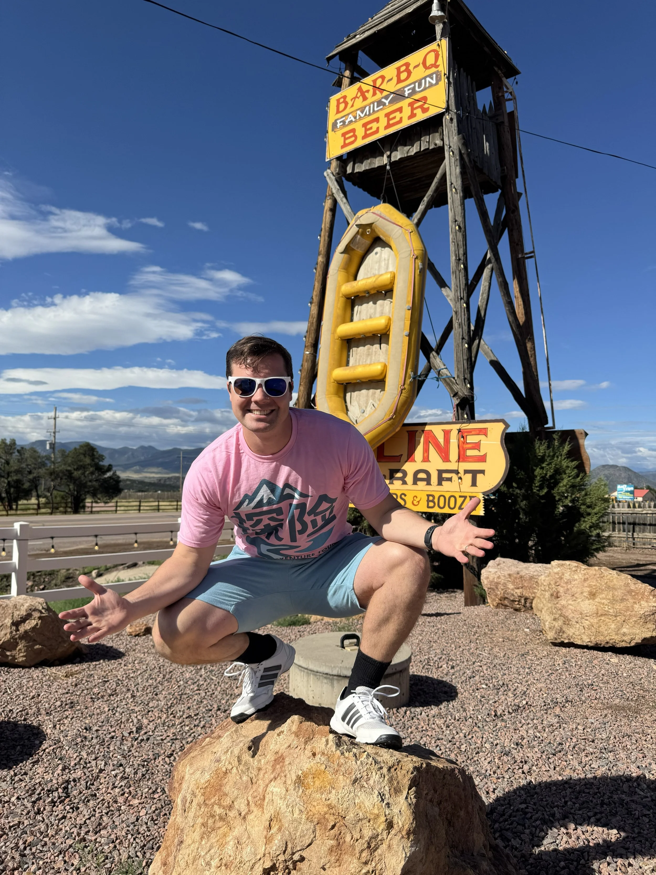 A man wearing sunglasses, a pink t-shirt, and light blue shorts crouches with arms open in front of a colorful sign that reads 'Bar-B-Q,' 'Family,' 'Fun,' 'Beer,' and 'Line Craft.' He is on a large rock outdoors under a partly cloudy sky.