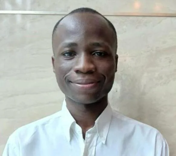 Headshot of a man smiling, wearing a white collared shirt, against a light-colored wall background.