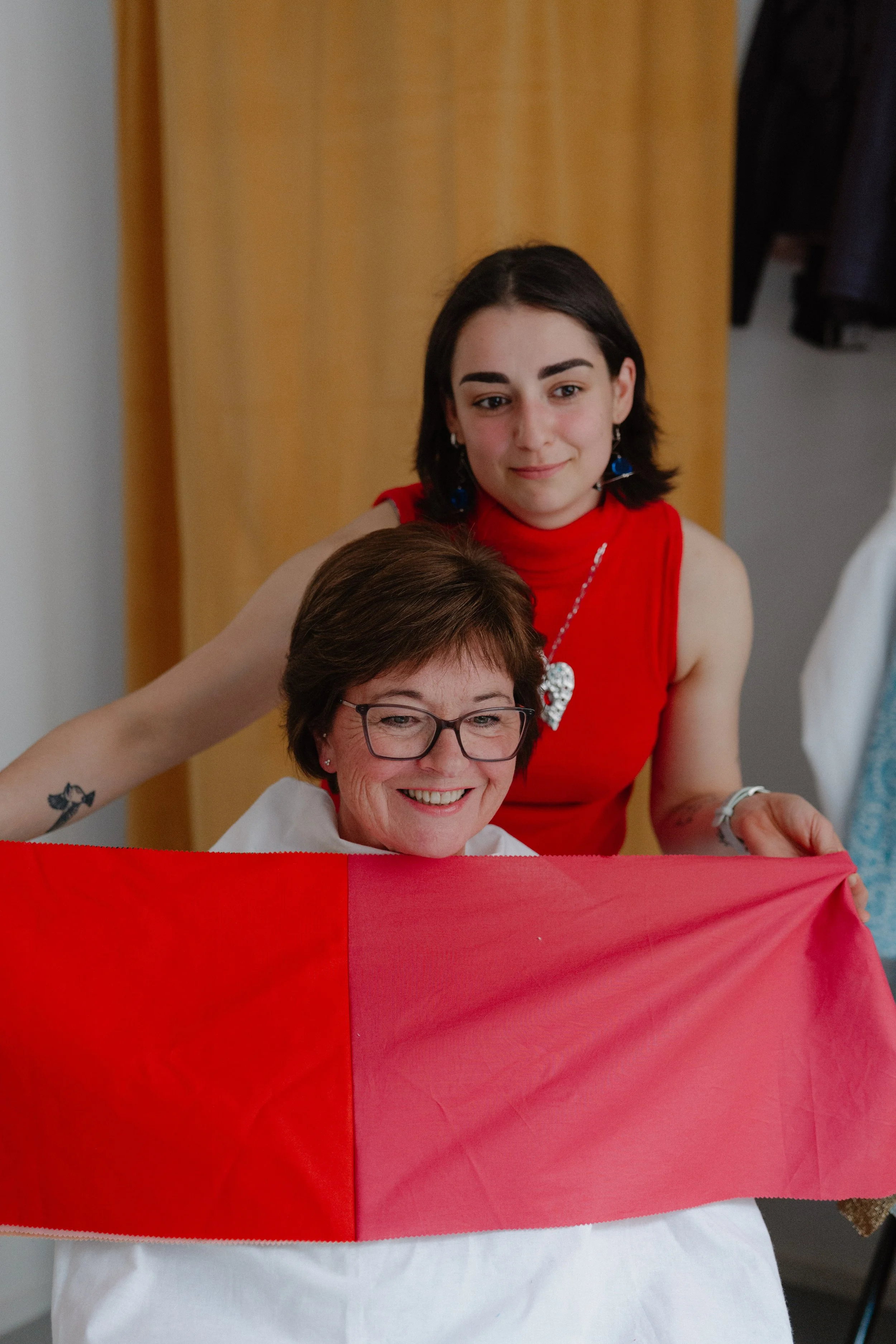 Two women, one older and wearing glasses, holding a red and white flag. The older woman is smiling, while the younger woman stands behind her, gently resting her hand on her shoulder.