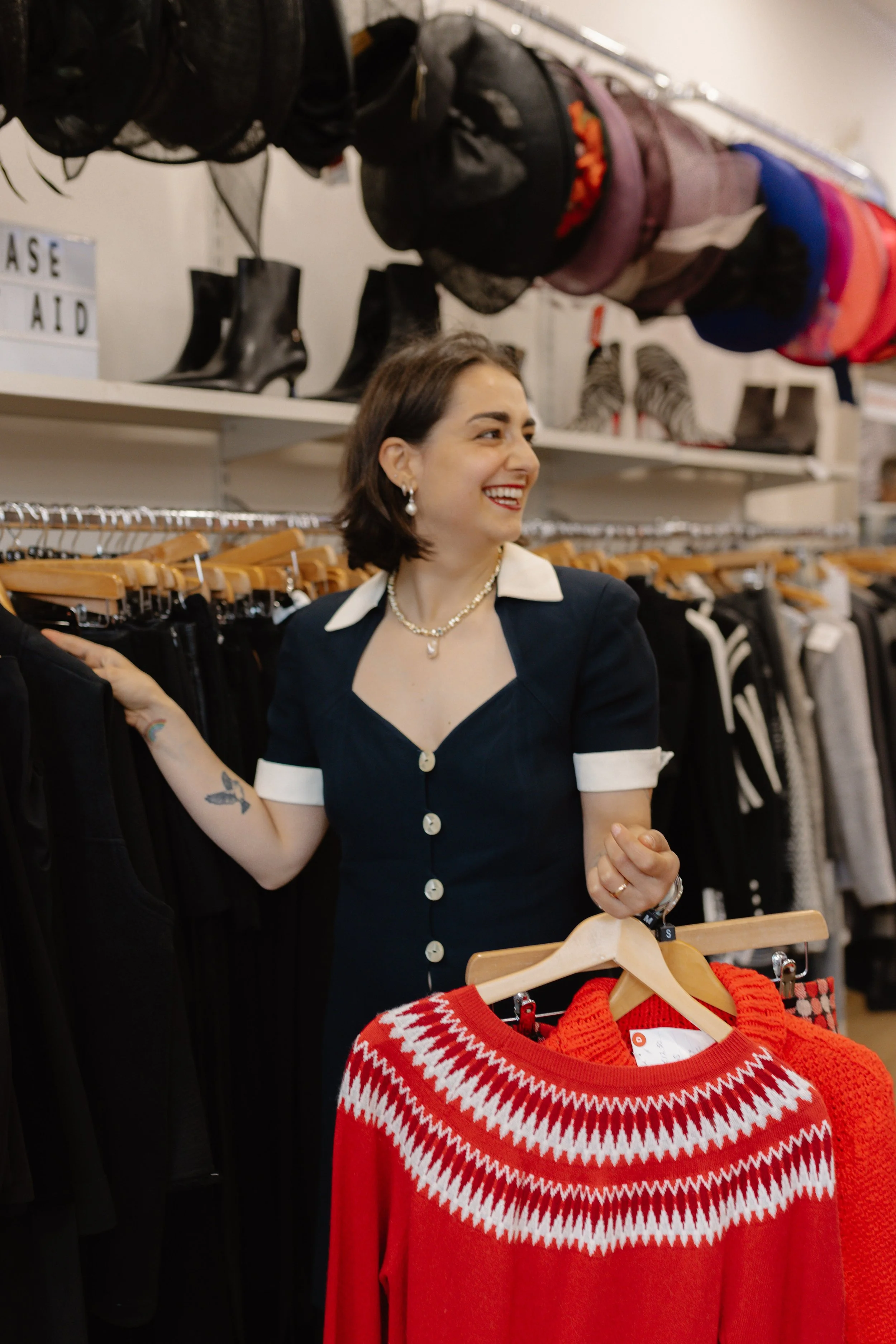 A woman smiling shopping in a clothing store, holding a red and white patterned sweater. She has short dark hair, wears jewelry, a black dress with white accents, and tattoos on her arm. The background shows racks of clothes, boots, and hats.
