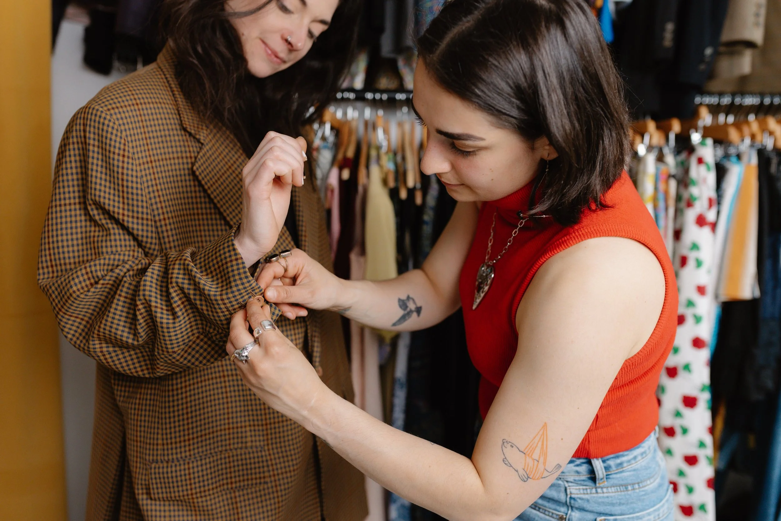 Two women are in a clothing store; one is trying on a brown checked blazer, while the other adjusts a ring on her finger. The woman in the blazer has dark hair and a nose ring. The other woman has a tattoo of a fish on her arm, short dark hair, and is wearing a red sleeveless top and jeans.