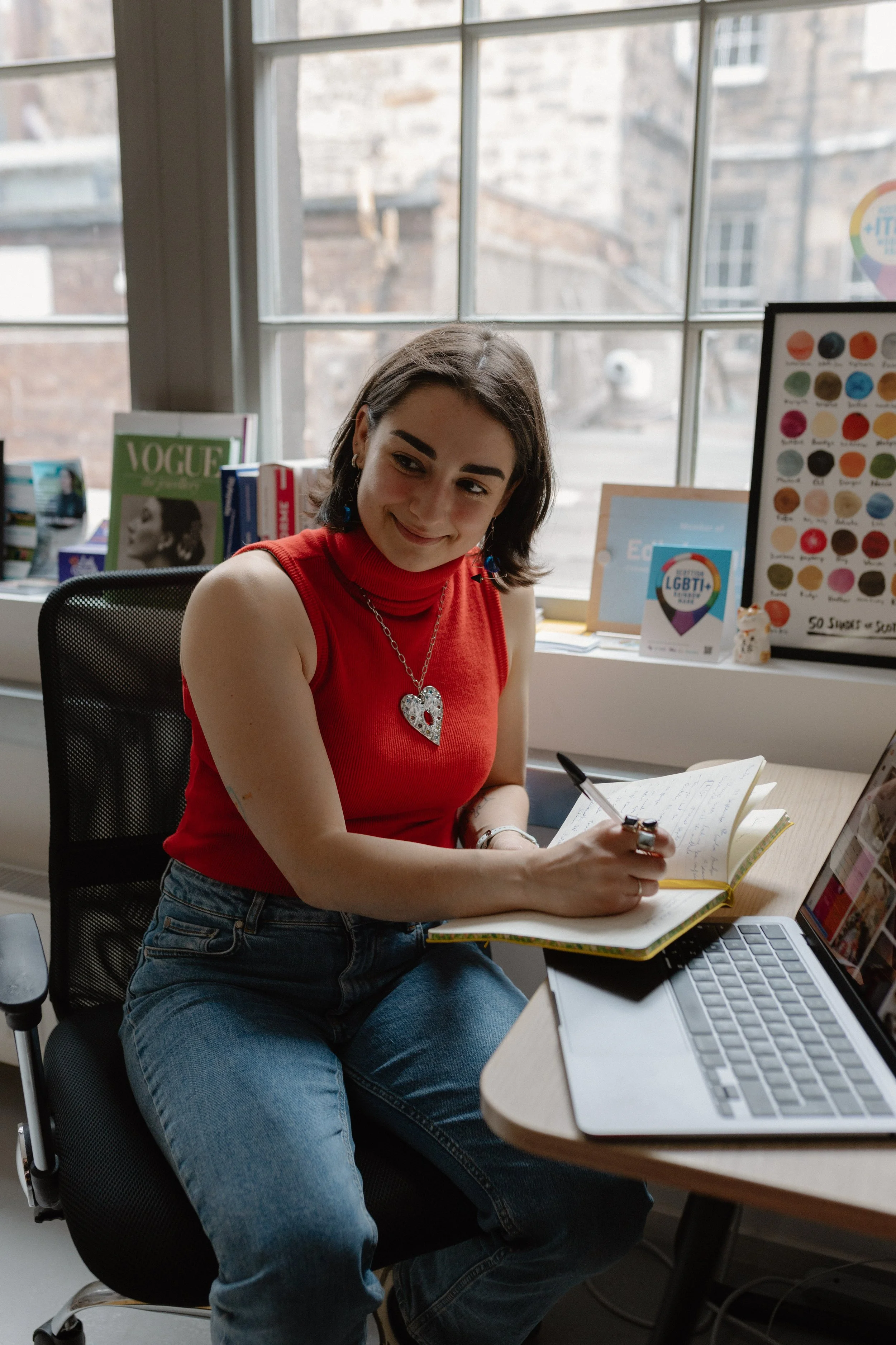 A young woman sitting at a desk, writing in a notebook, with a laptop open in front of her, in an office with large windows and colorful posters in the background.
