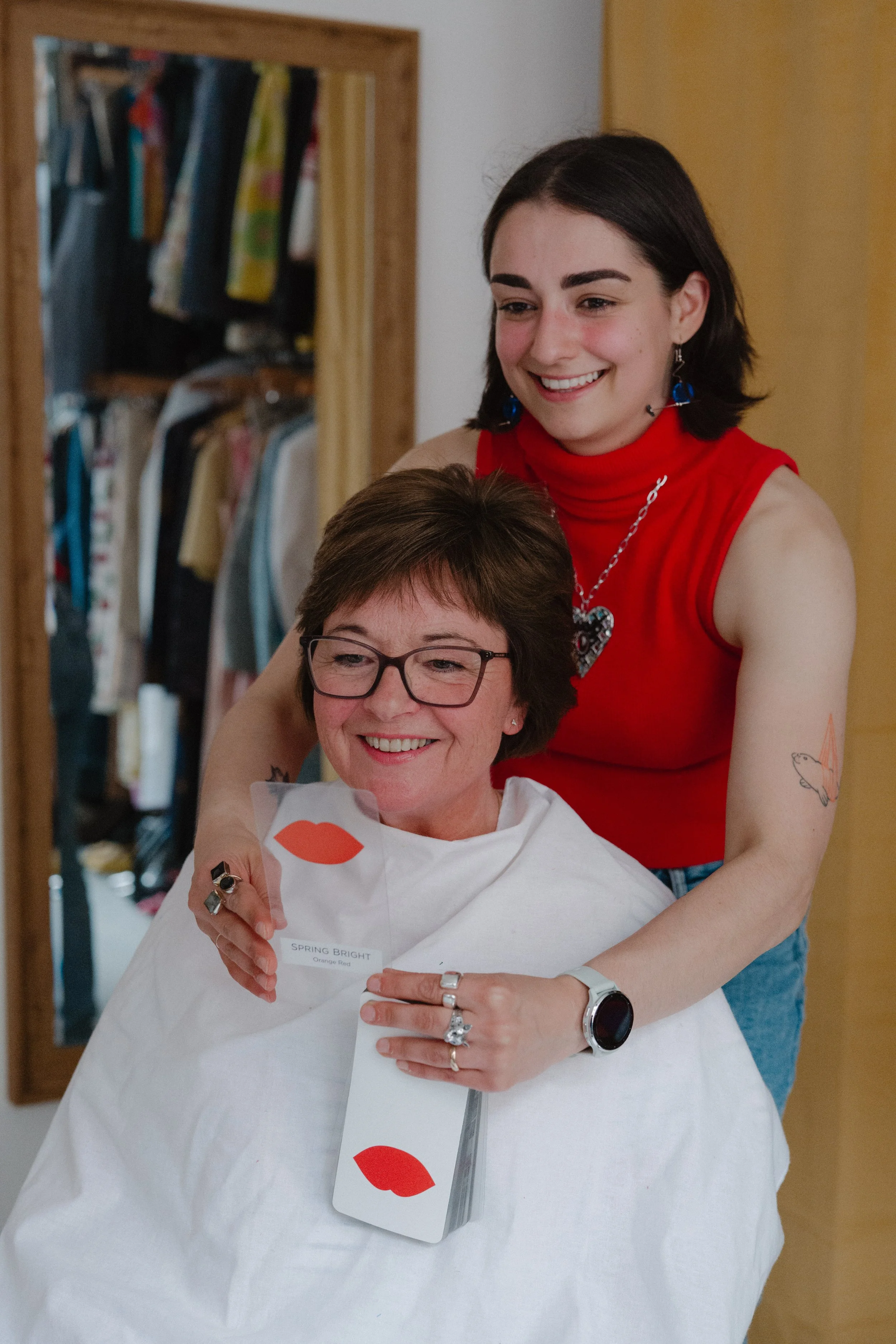 Two women, one older and one younger, are smiling while looking at a card with red lips. The older woman is seated with glasses and a white cape, and the younger woman is standing behind her wearing a red sleeveless top and jewelry. They appear to be in a room with a closet visible in the background.