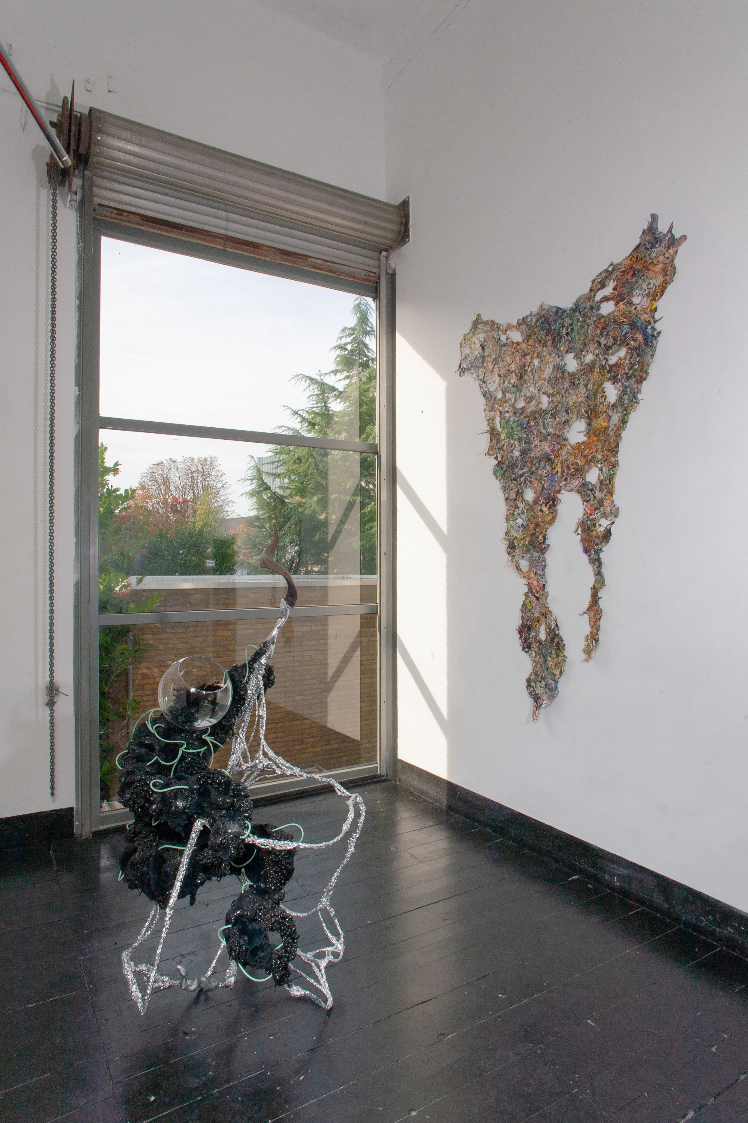 Contemporary art installation in a gallery featuring a large collage of materials forming the shape of a dog on the wall, a complex sculpture with black and silver materials on a black wooden floor, and a window showing trees outside.