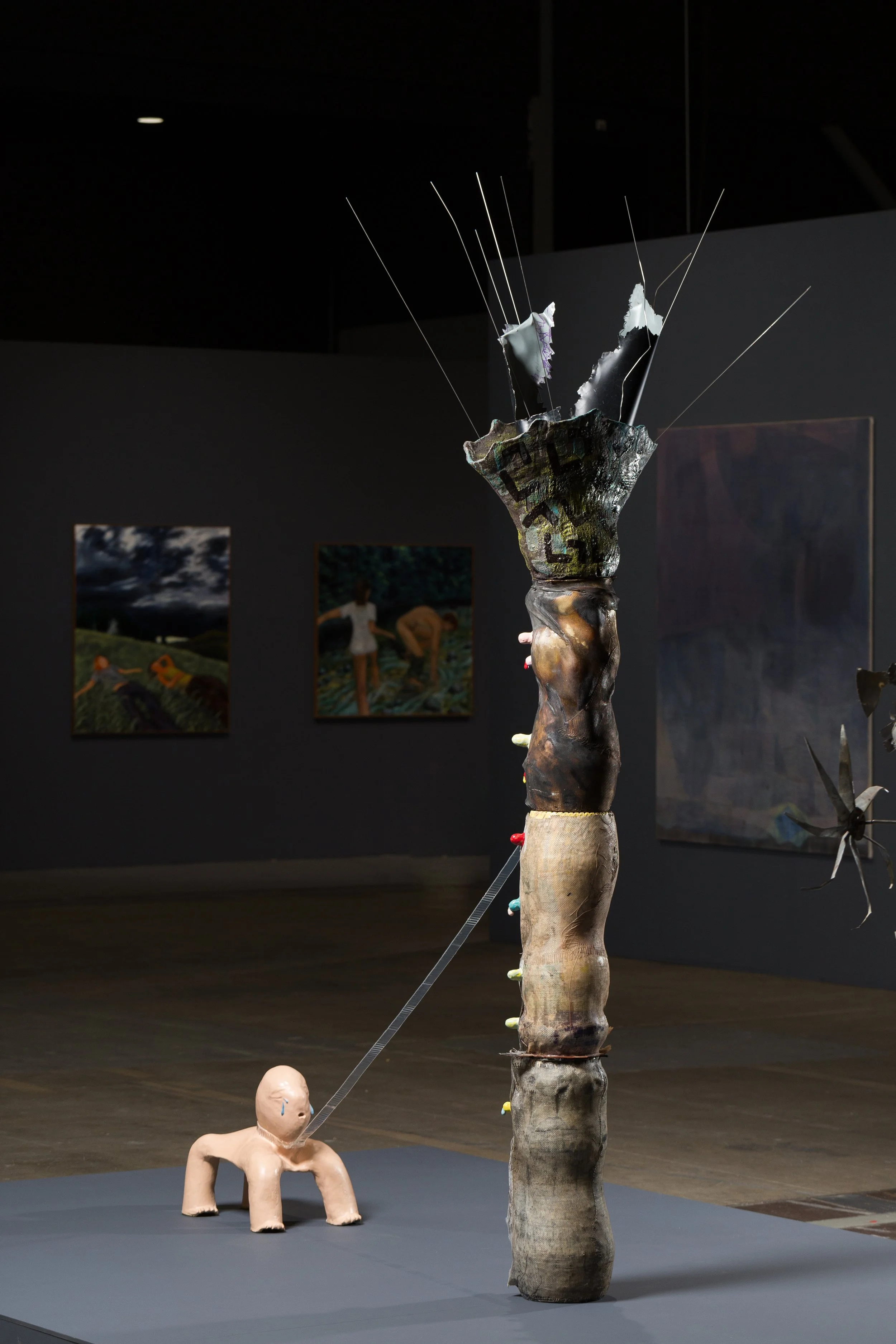 Mixed media art installation featuring a humanoid doll with a tear on its face, connected to a tall, abstract sculpture resembling a tree trunk with a textured surface, placed on a gray platform in an art gallery.