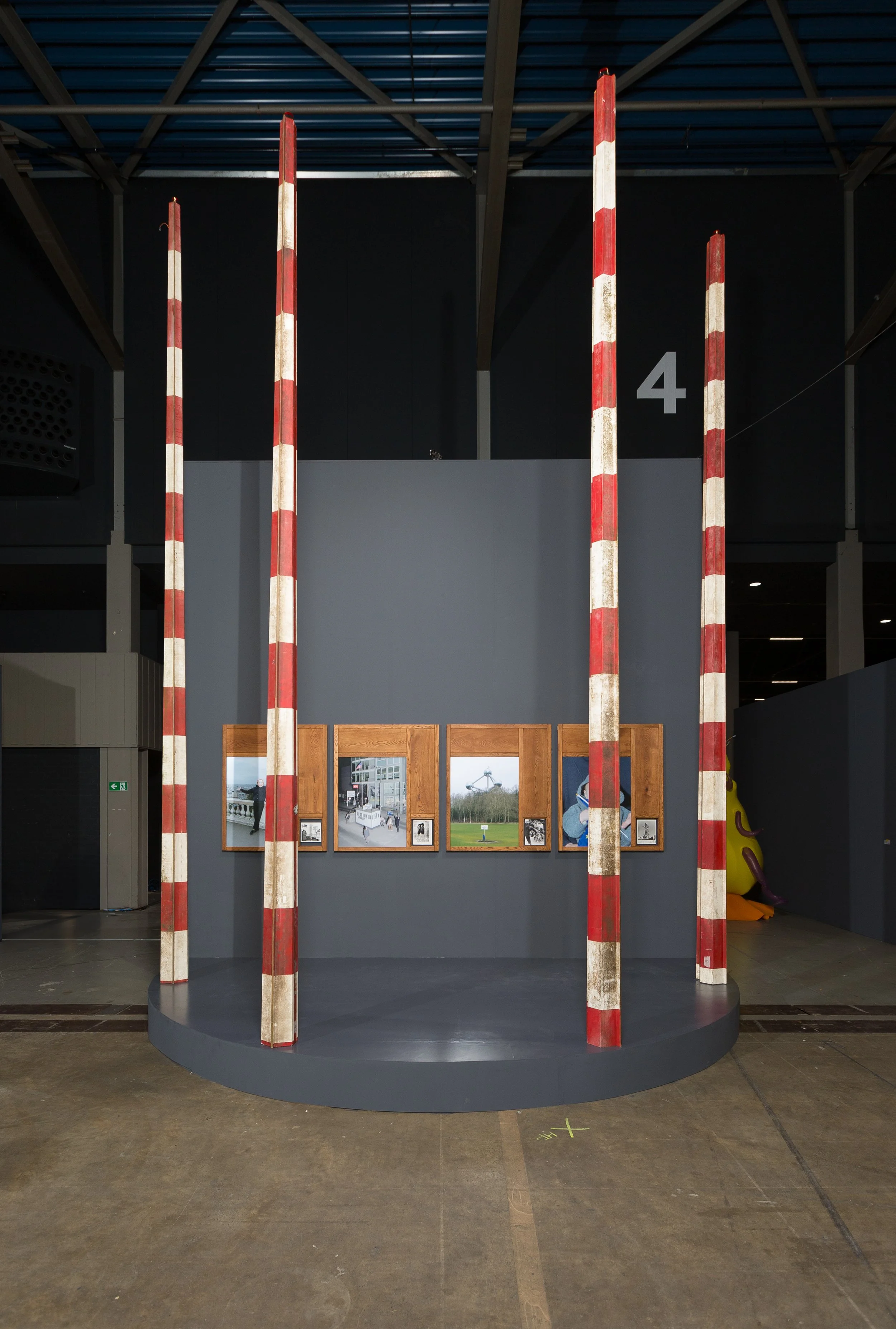 Indoor exhibition with four red and white striped poles standing in front of a display wall with photographs. A brown wooden door and a yellow inflatable object are partly visible on the right side.