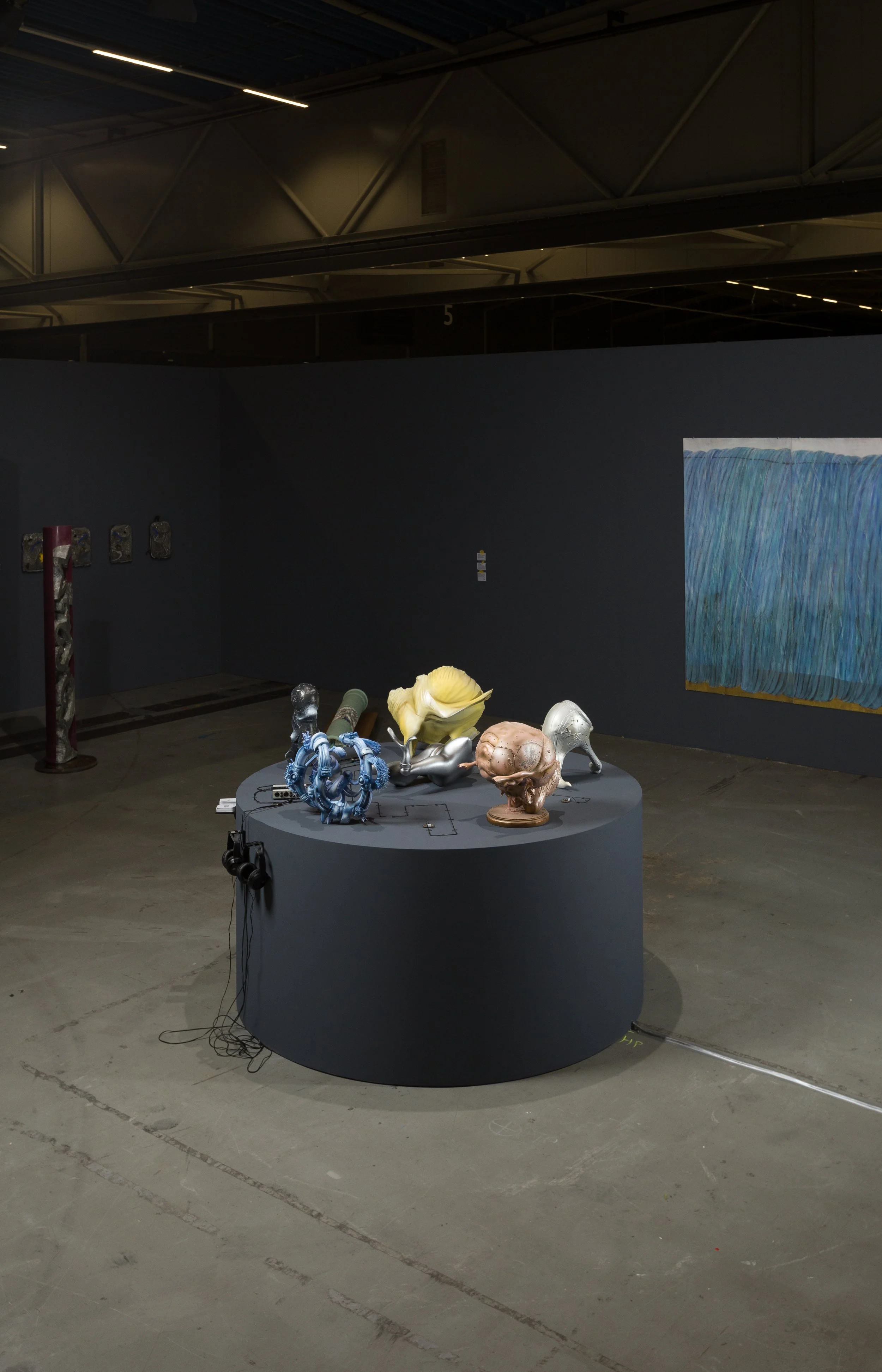 An art gallery with a dark gray circular display table featuring various sculptural objects, including a blue twisted sculpture, a yellow flower-like piece, and a skull. The background shows a black wall with artwork, and the gallery has a high industrial ceiling with exposed beams and lighting.