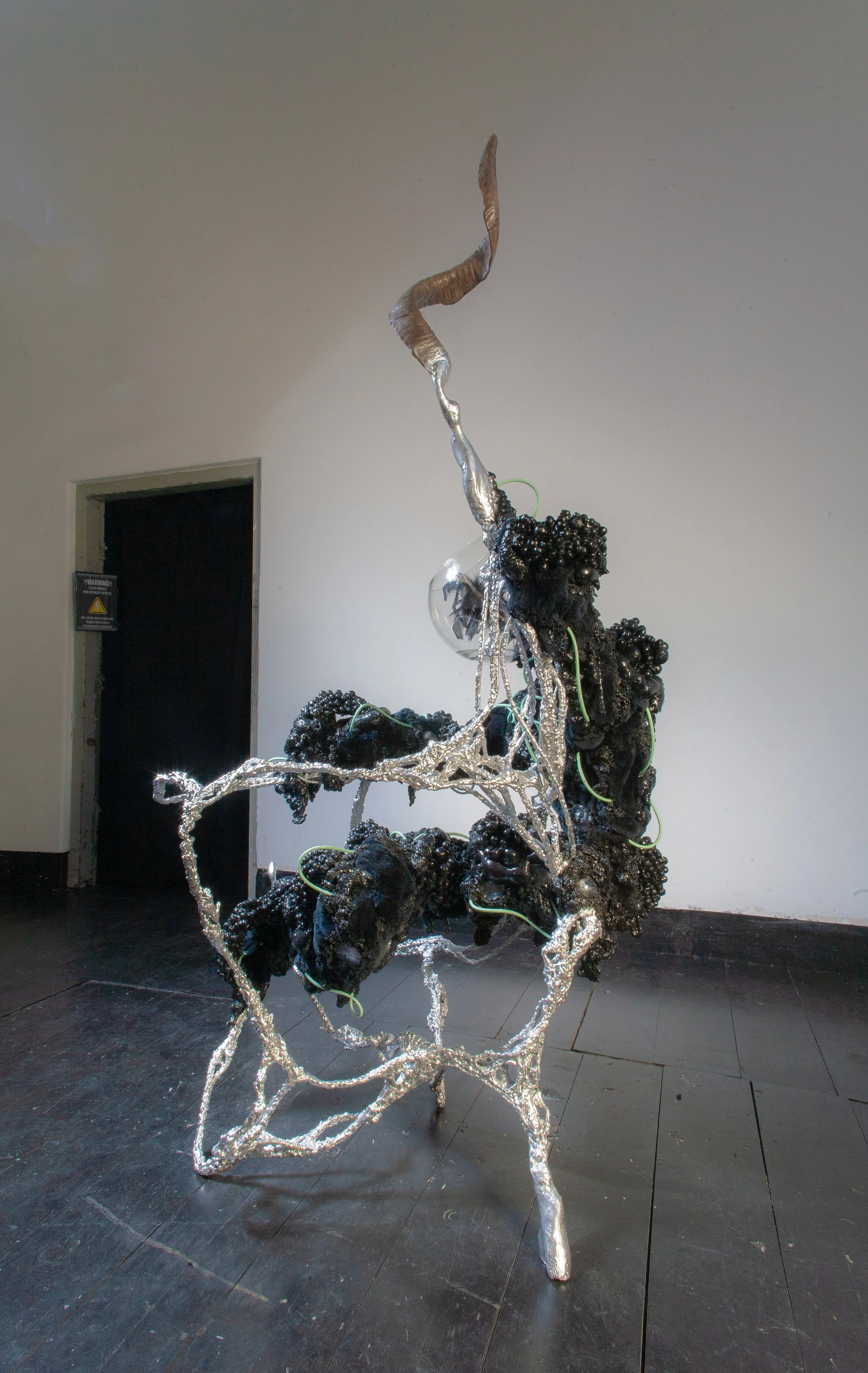 Contemporary sculpture on display in an art gallery; features a twisted metallic structure resembling a tree with black, irregularly shaped formations and green wires intertwined.