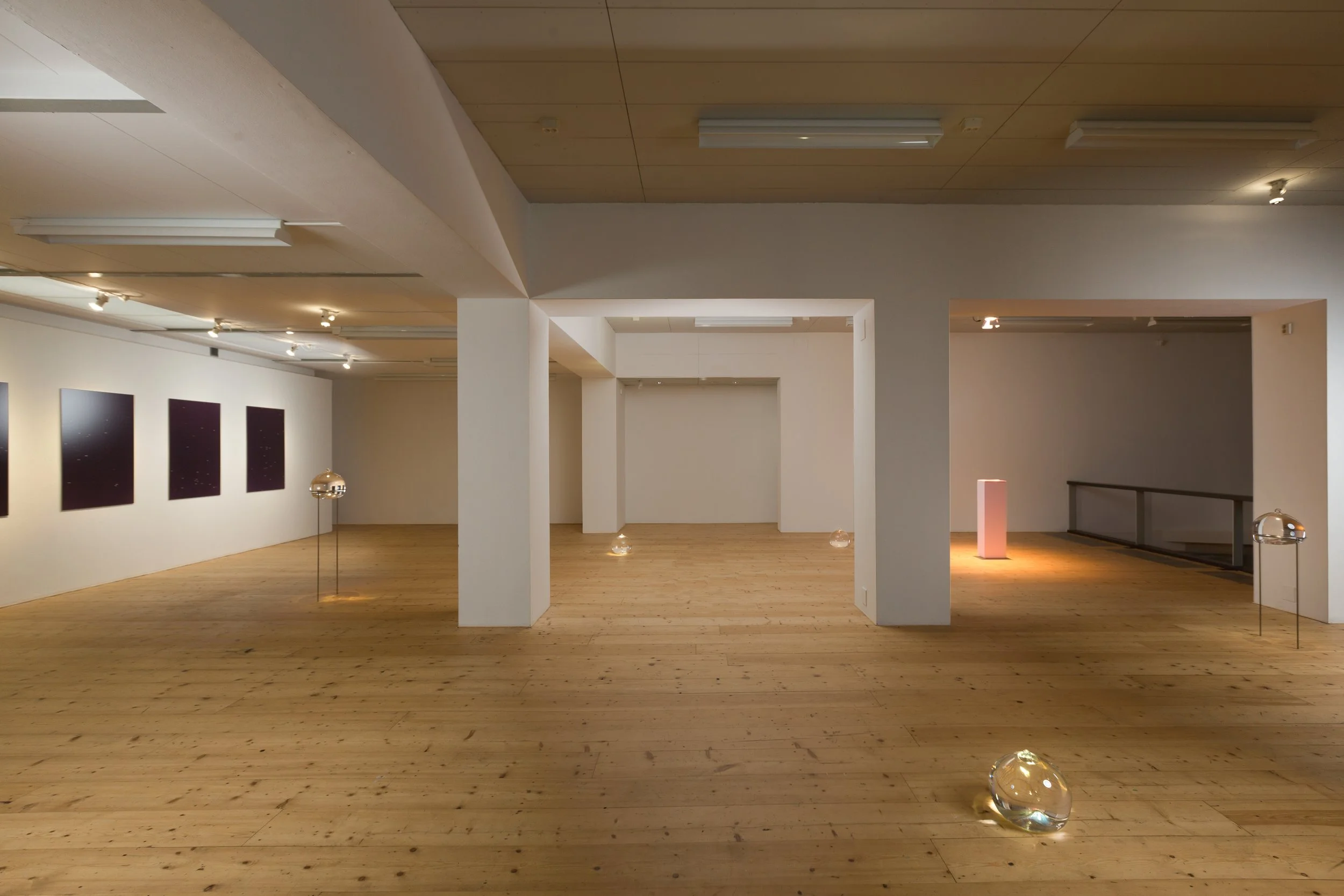 An art gallery featuring white walls with three framed pieces, a wooden floor, and spotlights on the ceiling, with small glass sculptures on the floor.